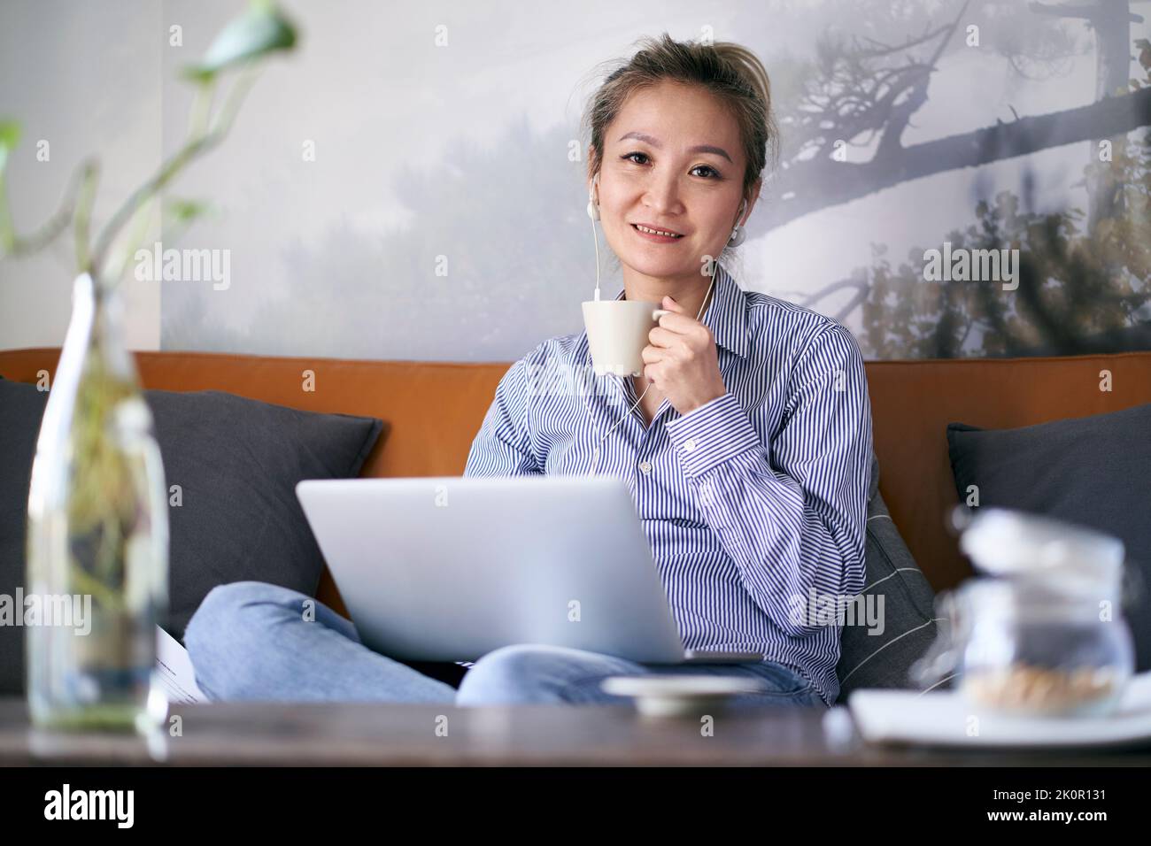 Reife professionelle asiatische Frau, die von zu Hause aus auf der Couch sitzt, mit einem Laptop, der eine Tasse Kaffee hält und die Kamera anschaut Stockfoto