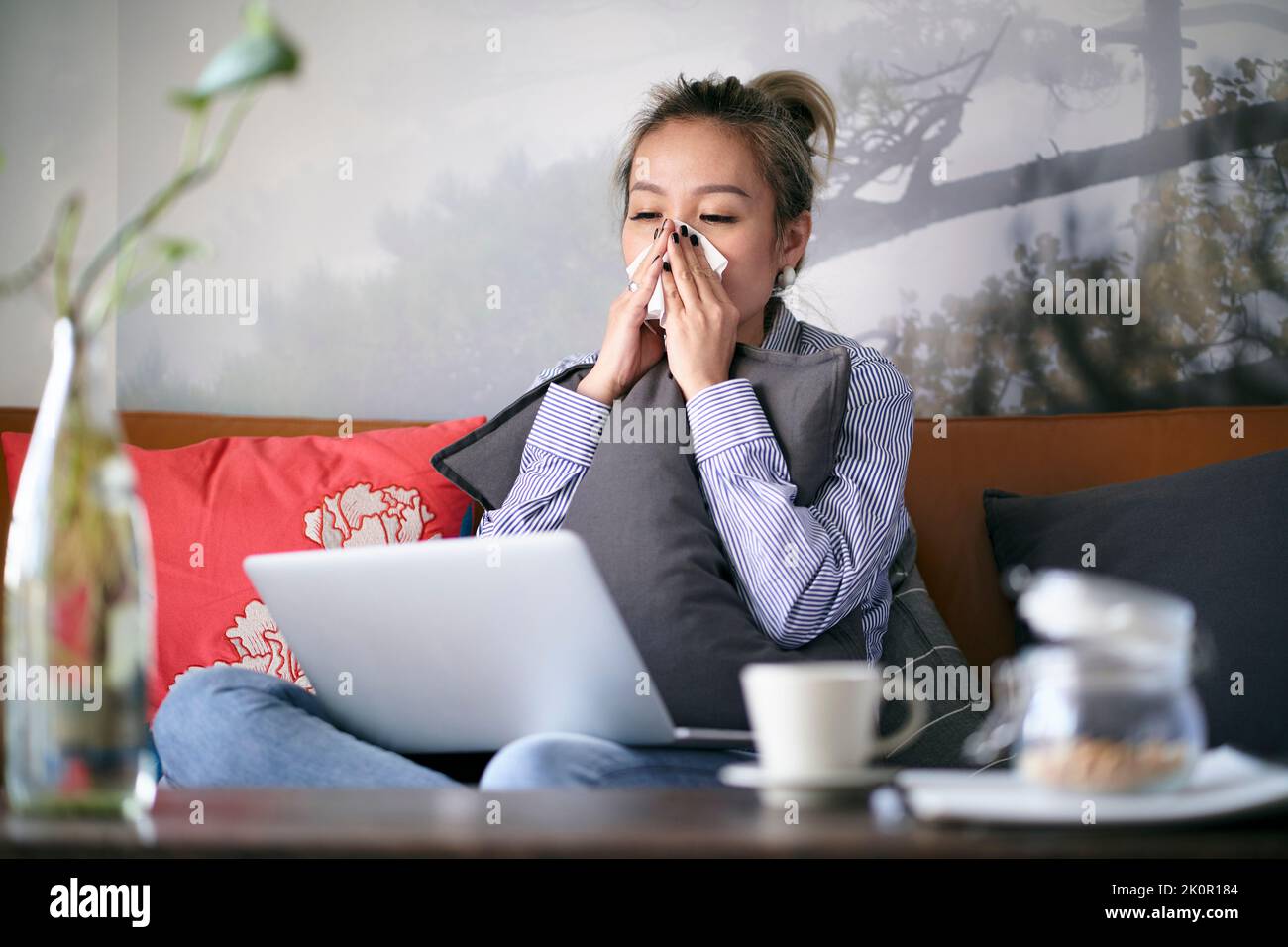 Reife asiatische Frau, die sich erkältet, mit laufender Nase und von zu Hause aus mit einem Notebook auf dem Sofa sitzt Stockfoto