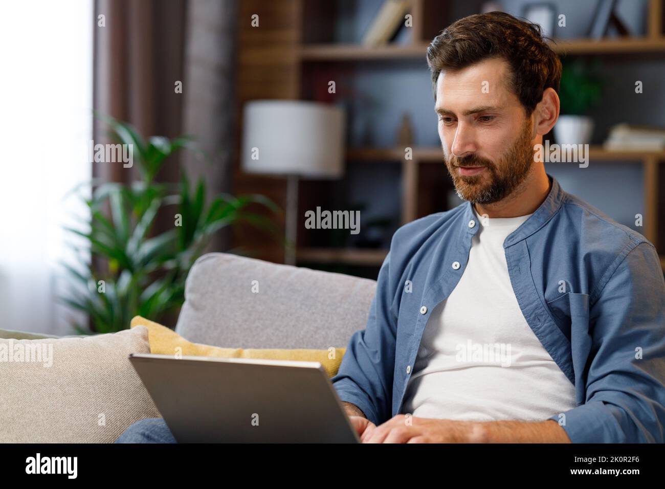 Ein hübscher Mann, der in einer stilvollen Wohnung auf dem Sofa sitzt und am Laptop arbeitet. Arbeiten im Internet, Nutzung sozialer Netzwerke, Unterhaltung in der Wohnung Stockfoto