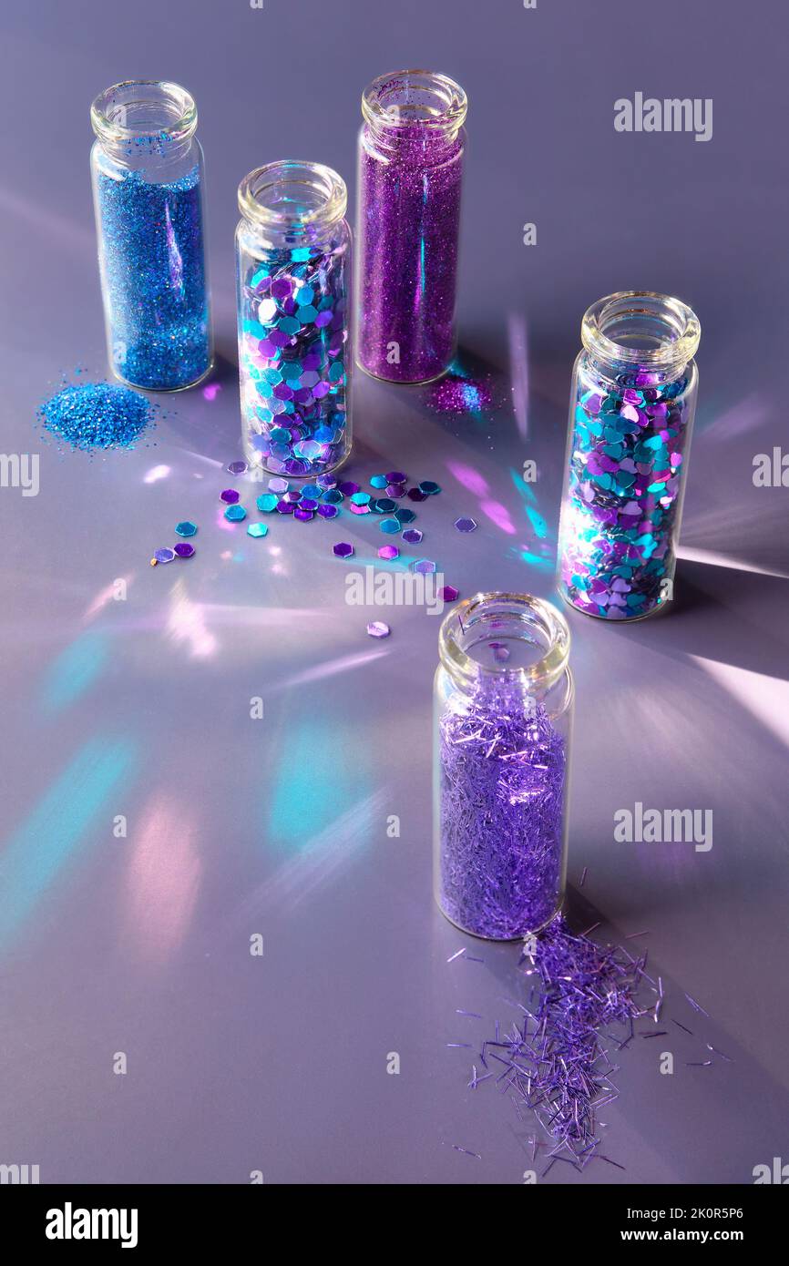 Glitzer-Produkte in transparenten Flaschen, Nahaufnahme. Verschiedene Glitzerpulver und Partikel in neonblauen, violetten und türkisfarbenen Tönen auf dunklem Hintergrund. Stockfoto