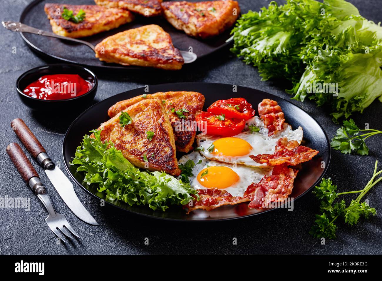 irisches Frühstück mit Spiegeleiern, Speckscheiben, gebratenen Kartoffelkuchen, gegrillten Tomaten und frischem Salat auf schwarzem Teller auf Betontisch mit Zutaten Stockfoto