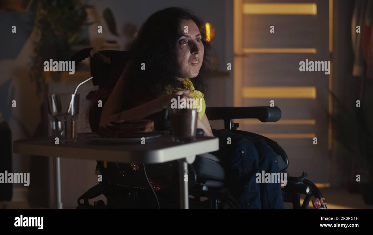 Frau mit körperlicher Behinderung sitzt im motorisierten Rollstuhl vor dem Fernseher und entspannt sich zu Hause in der Nacht beim Fernsehen Stockfoto