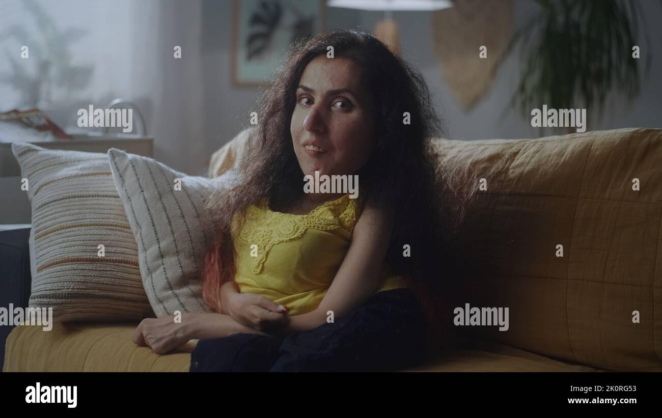 Attraktive Frau mit körperlicher Behinderung lächelt und hebt ihre Augenbraue, während sie zu Hause auf dem Sofa sitzt und die Kamera anschaut Stockfoto