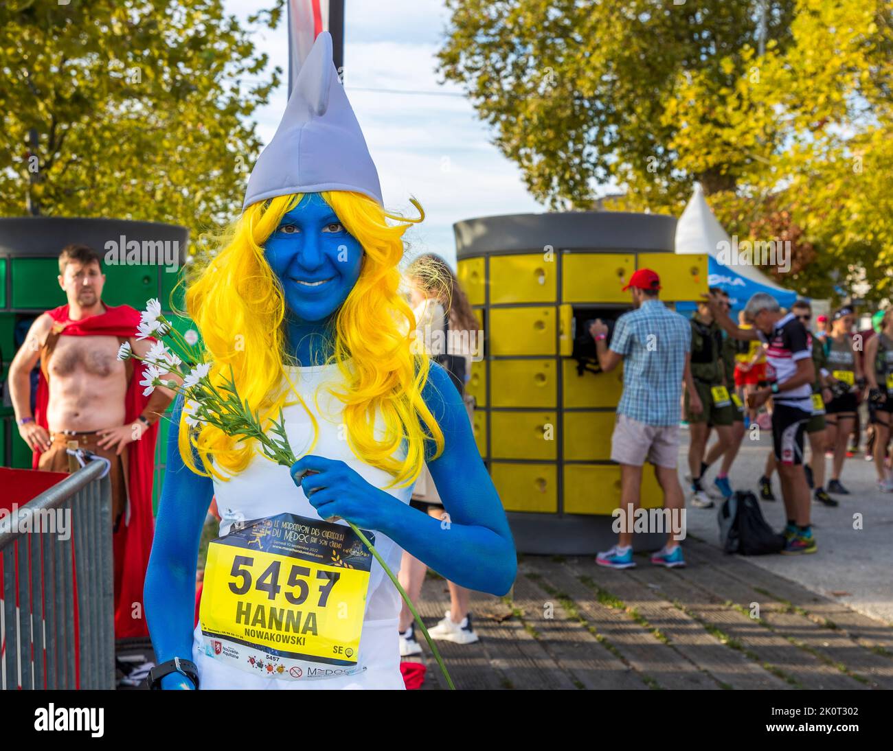 Ein Läufer, der blau angemalt und als Smurfette verkleidet ist, trägt eine weiße spitze Mütze und eine gelbe Perücke und posiert mit Startnummer 5457 während der Kostümparade des 36. Marathon des Châteaux du Médoc am Quai Léon Perrier in Pauillac, Frankreich. Das Thema 2022 ist „Film". Stockfoto