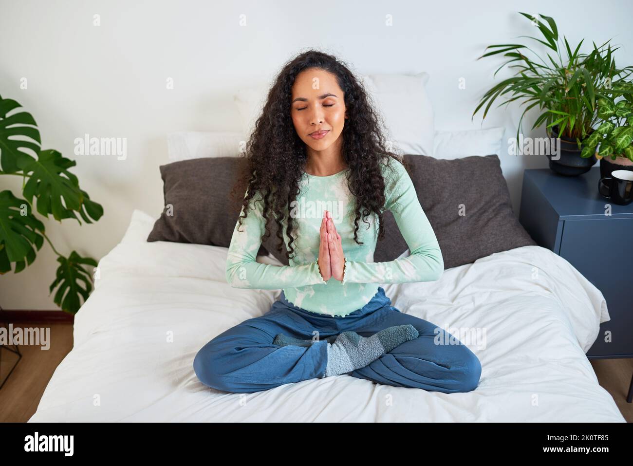 Eine junge, multiethnische Frau sitzt mit gekreuzten Beinen auf dem Bett und den Händen in Gebetshaltung Stockfoto