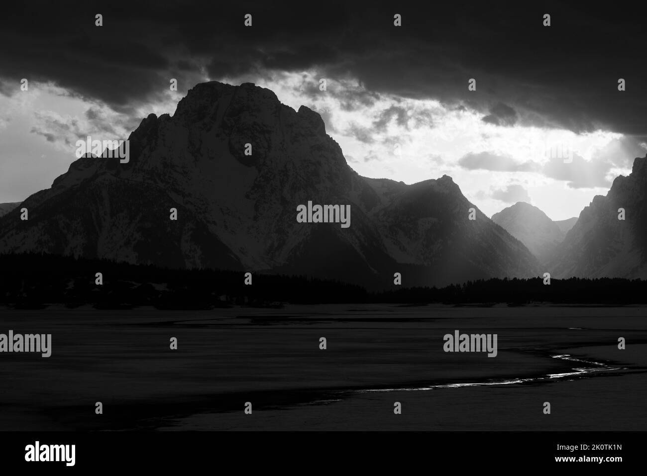 Eine untergehende Sonne verdunkelt sich hinter dichten Sturmwolken, die die Ufer des Jackson Lake erleuchten, wo das Eis aus der Wintersaison unter dem Mount Mora geschmolzen ist Stockfoto