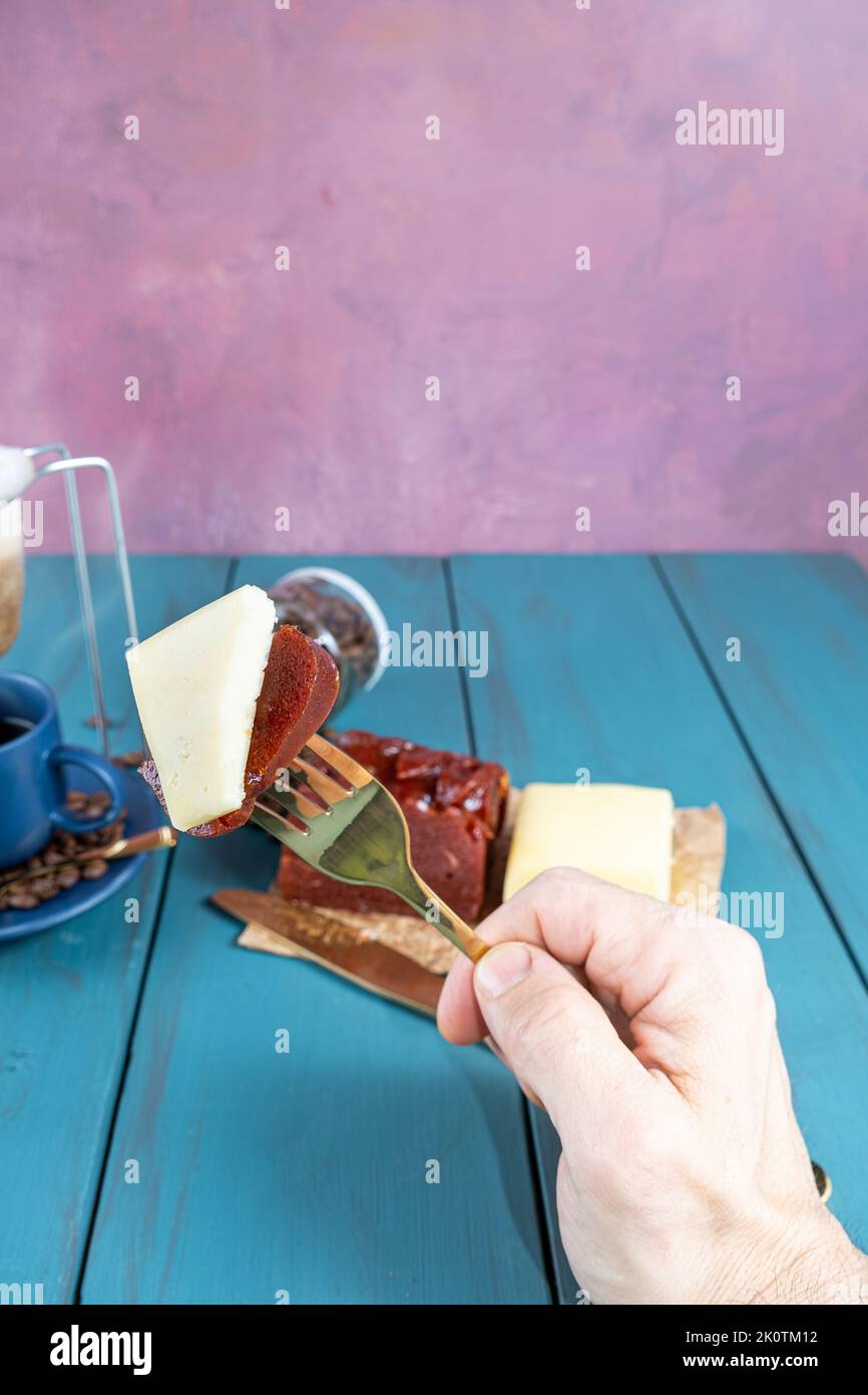 Mann, der eine Gabel mit Guava-Stücken mit Käse hält. Im unscharfen Hintergrund Bohnen und Kaffeetasse vertikal. Stockfoto