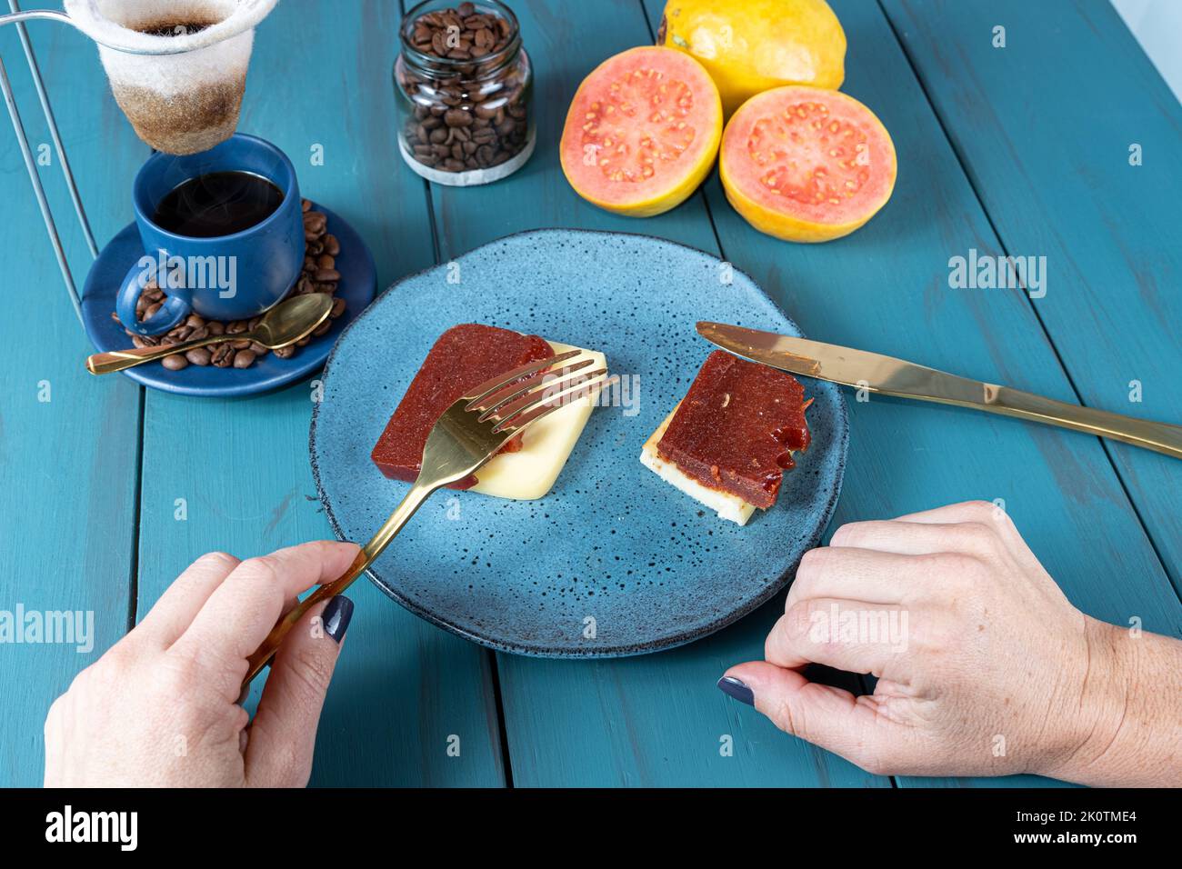 Frau, die eine Gabel über Guava-Scheiben mit Käse hält, umgeben von Bohnen und einer Tasse Kaffee. Stockfoto