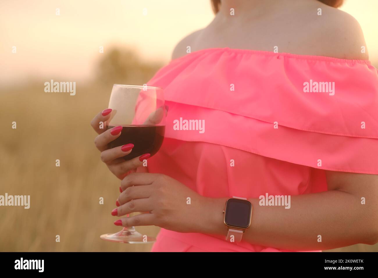 Nicht erkennbare Frau, die ein Glas Rotwein in der Hand hält und am Sonnenuntergang in der Natur steht. Körperteil des Weibchens in hellem Kleid mit Alkohol auf dem Land Stockfoto