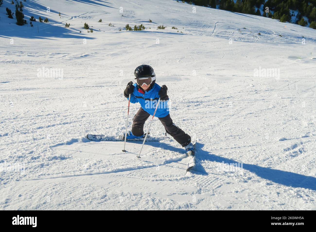 Ein kleiner Anfänger Skifahrer auf einer Skipiste in einer lustigen umgekehrten Position und lächelt hinter der Brille und schaut auf die Kamera, Kind hat Spaß beim Lernen Stockfoto
