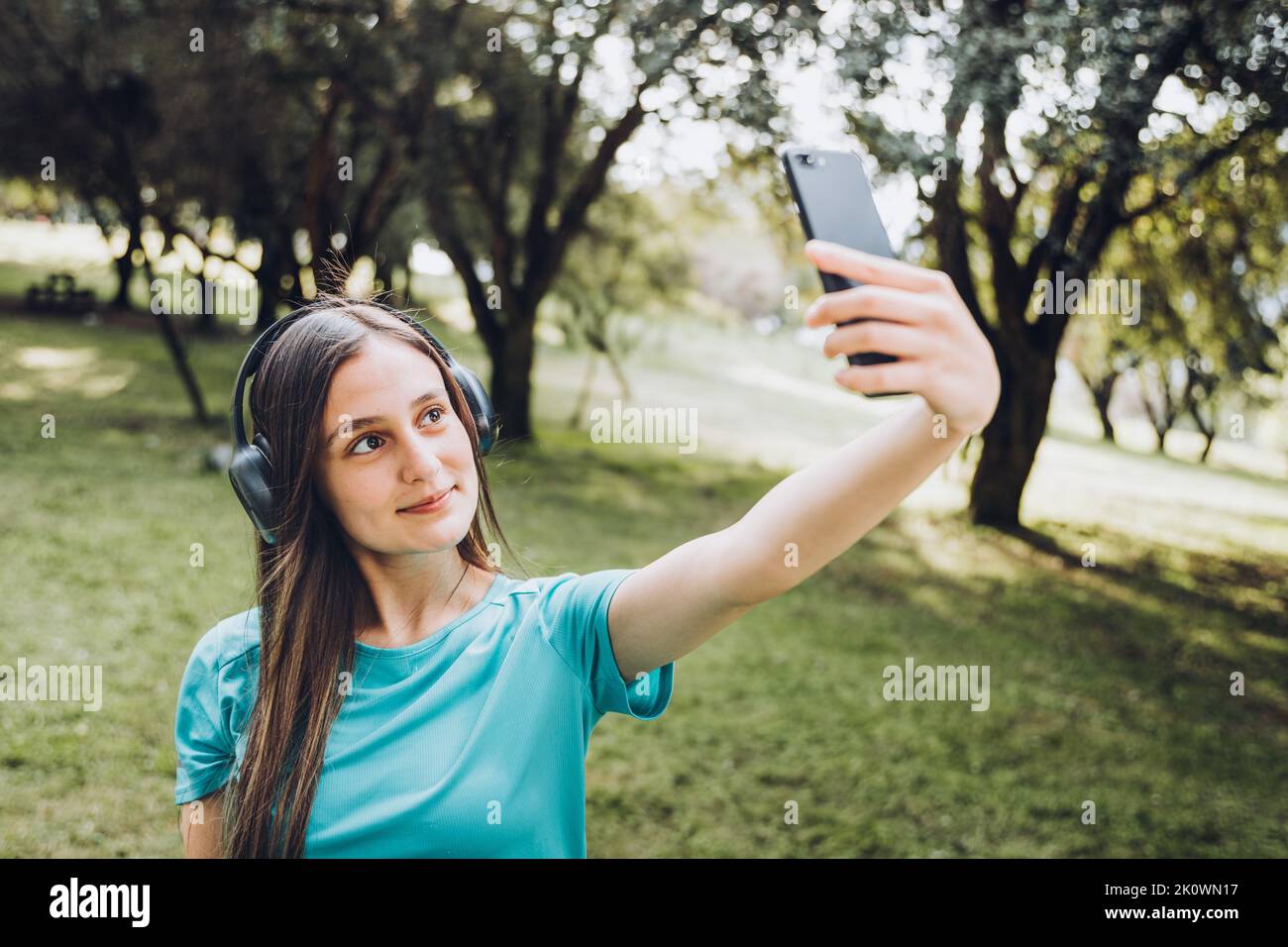 Lächelndes junges Mädchen, das ein türkisfarbenes T-Shirt und Kopfhörer trägt und ein Selfie mit ihrem Handy in einem natürlichen Raum macht Stockfoto