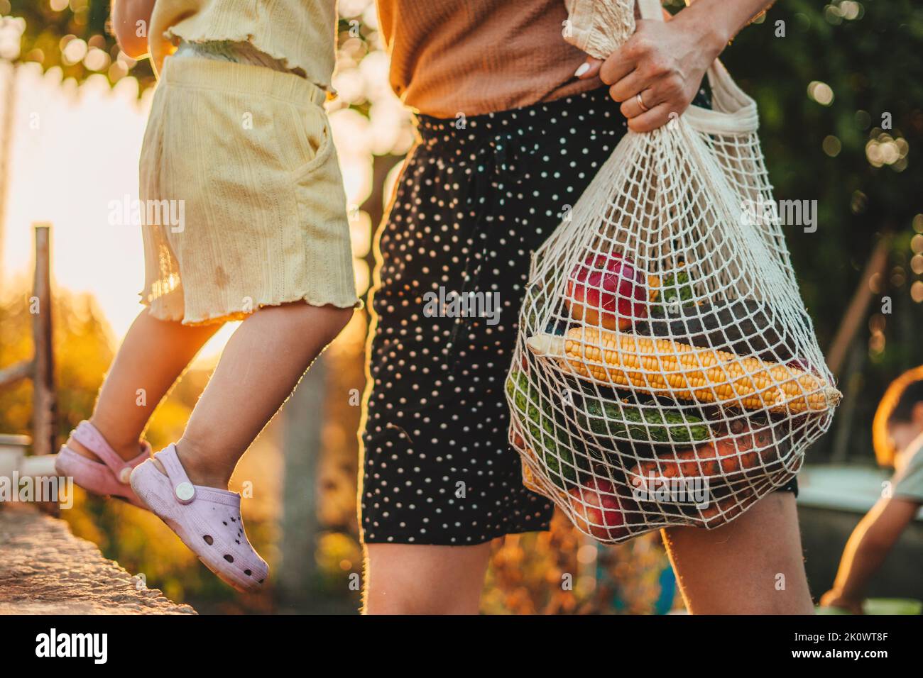 Beschnittene Ansicht der Hand einer Farmerin, die einen Öko-Beutel mit frisch gepflücktem Gemüse und ein Baby in den Armen hält. Frisches Bio-Gemüse Stockfoto