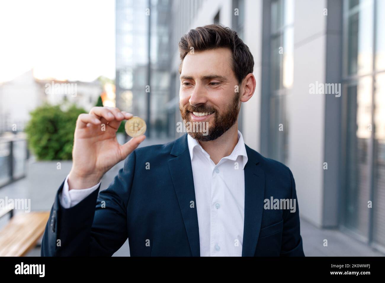 Froh zuversichtlich attraktiven jungen kaukasischen Kerl mit Bart im Anzug zeigt Bitcoin Münze im Freien Stockfoto