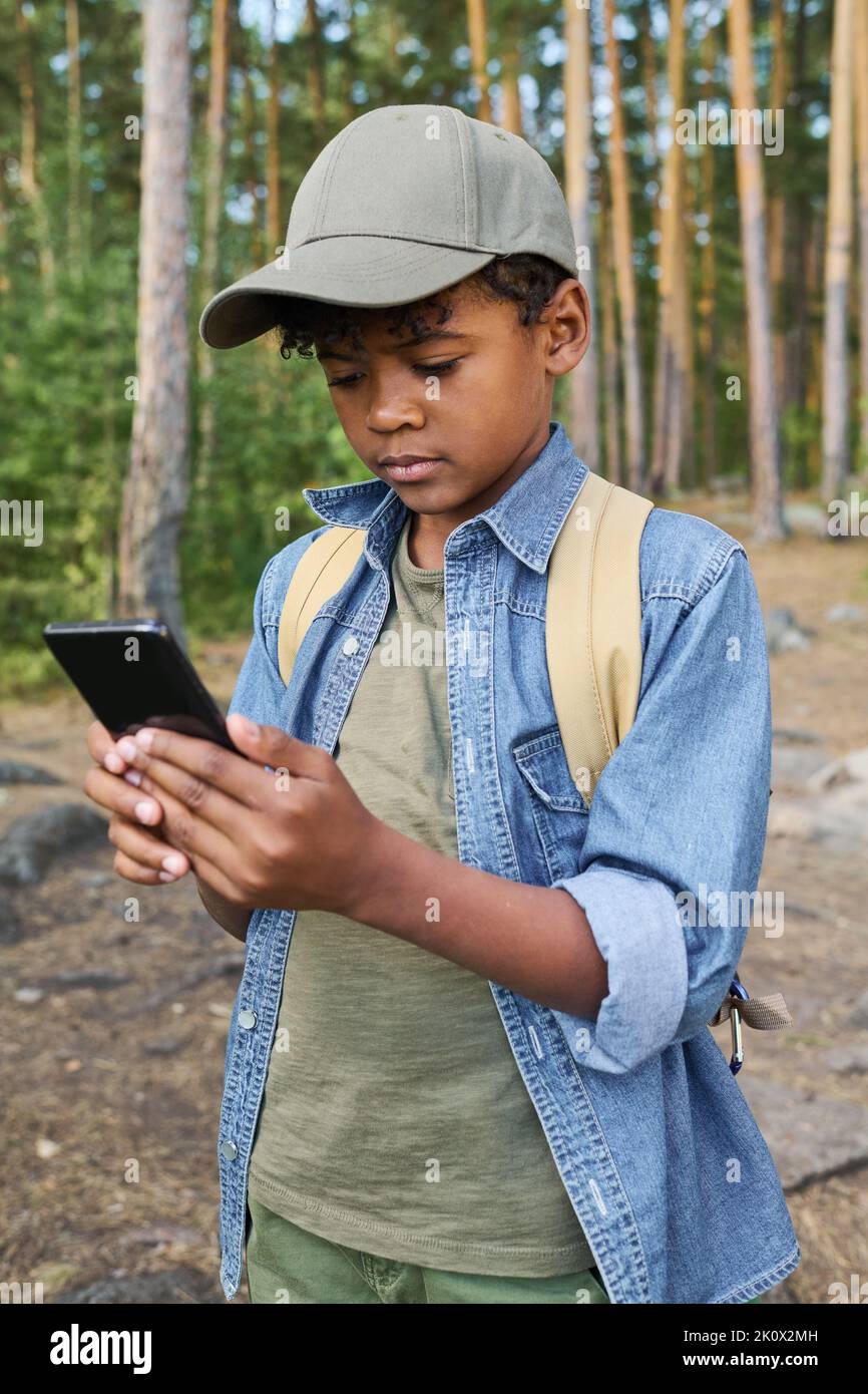 Jugendlicher afroamerikanischer Junge mit Smartphone und Navigatorkarte, während er während seiner Rucksackreise in tiefen Wäldern auf dem Weg steht Stockfoto