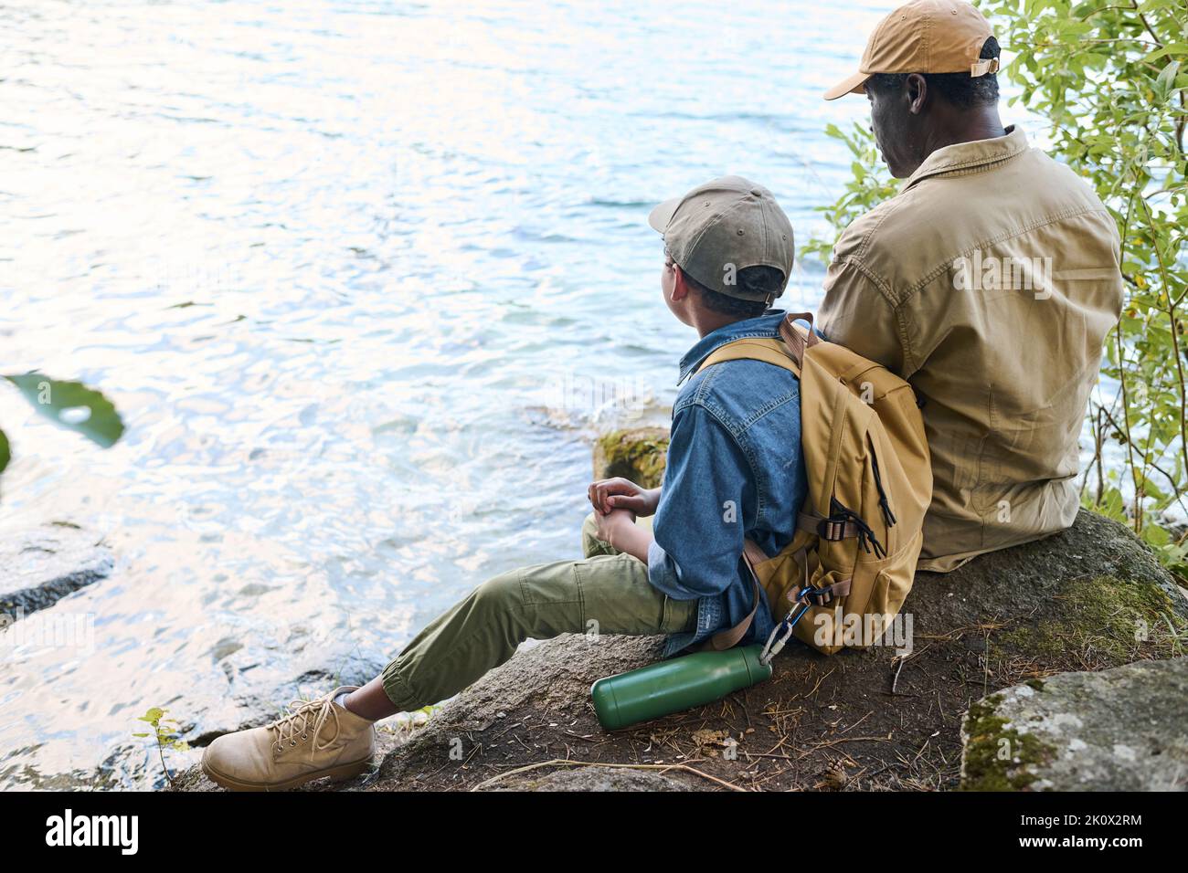 Rückansicht des afroamerikanischen Jungen mit Rucksack und seines Großvaters, der am Wasser sitzt und sich während der Wochenendwanderung ausruhen kann Stockfoto