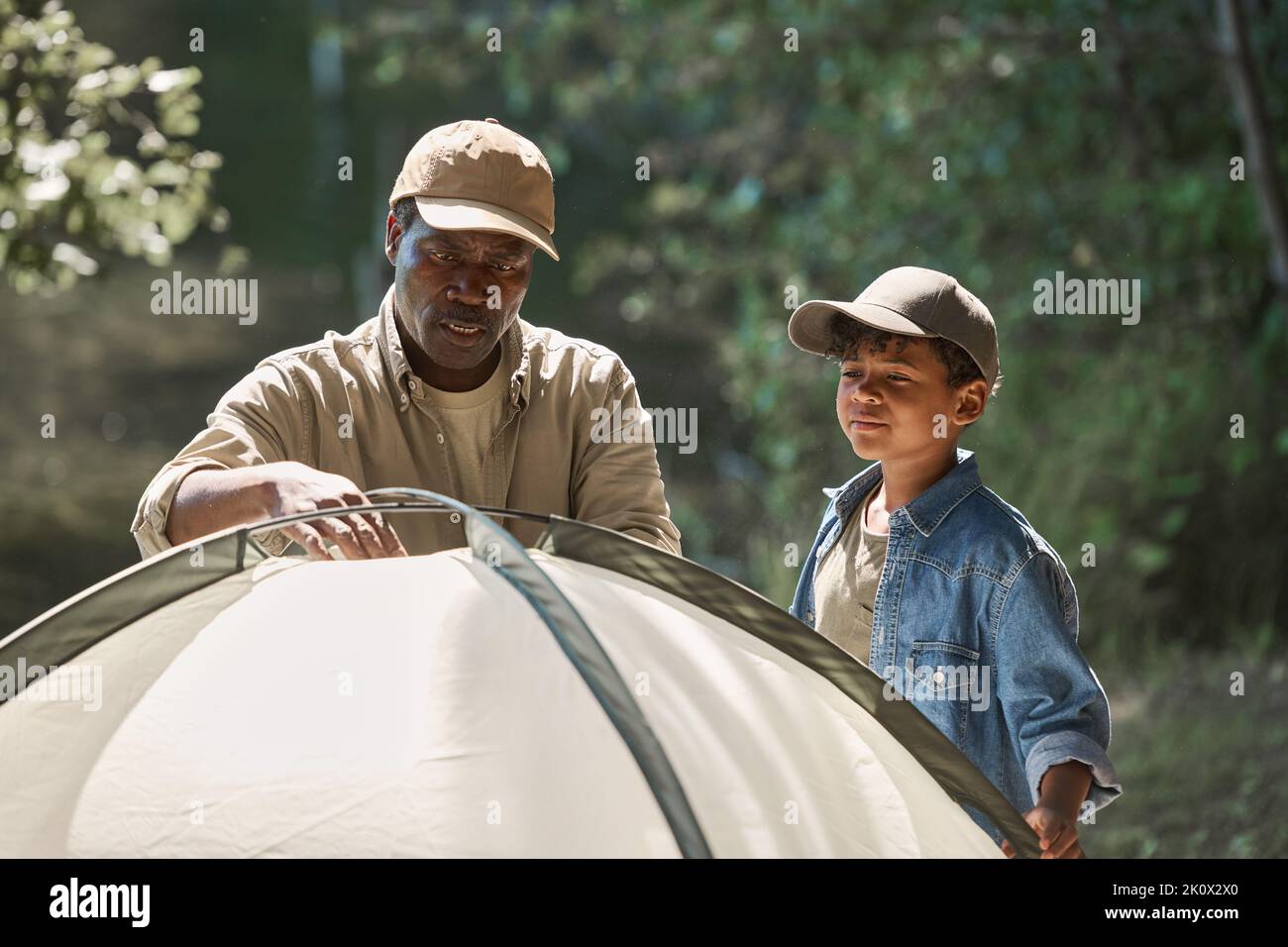 Ein süßer afroamerikanischer Junge, der seinem Großvater dabei hilft, Zelt im Wald zu schlagen, während beide am sonnigen Sommerwochenende ihren Wanderausflug genießen Stockfoto