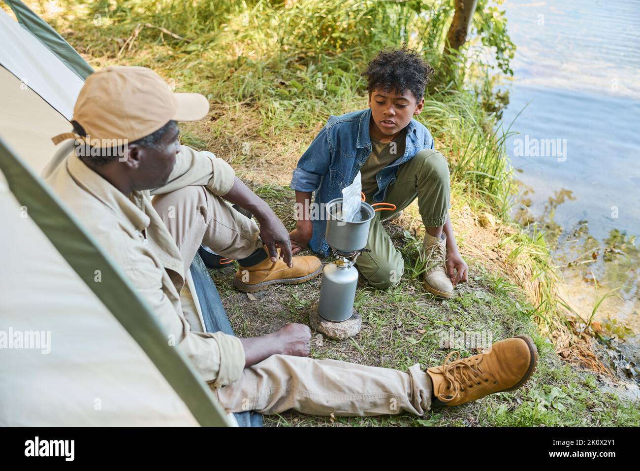 Jugendlicher afroamerikanischer Junge in Casualwear, der mit seinem Großvater spricht, während er am Wasser sitzt und darauf wartet, dass das Essen gekocht wird Stockfoto