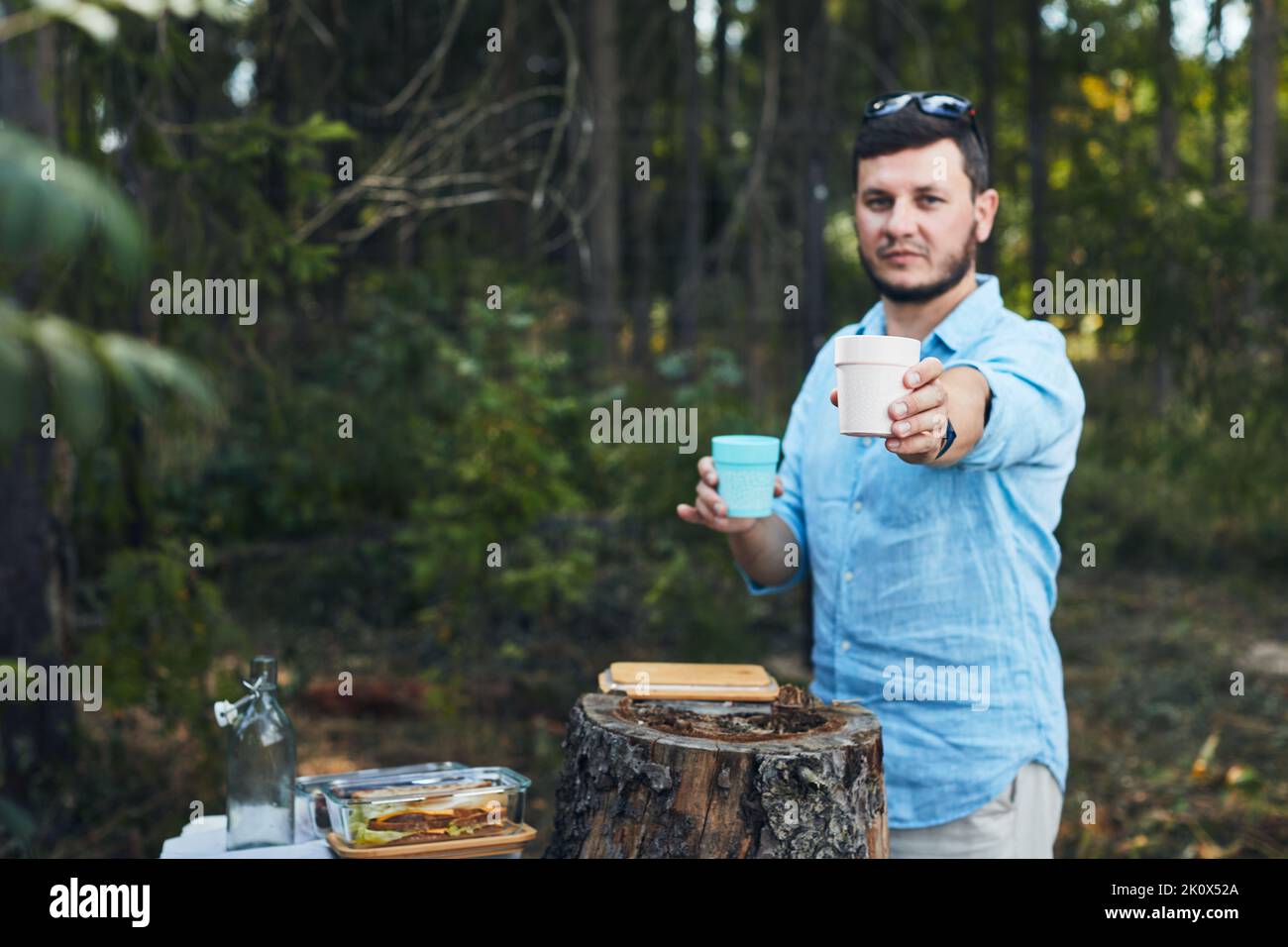 Ein junger Mann bei einem Picknick reicht ein Glas mit einem Getränk zur Kamera. Vorderansicht. Stockfoto