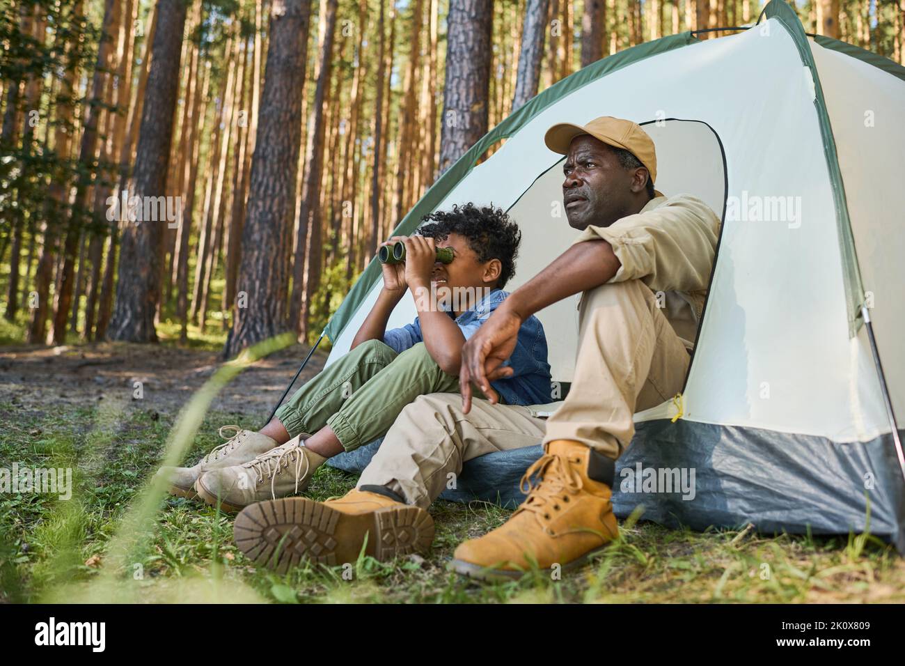 Süßer afroamerikanischer Junge, der durch ein Fernglas schaut, während er im Zelt neben seinem Großvater in Casualwear während der Wanderung im Wald sitzt Stockfoto