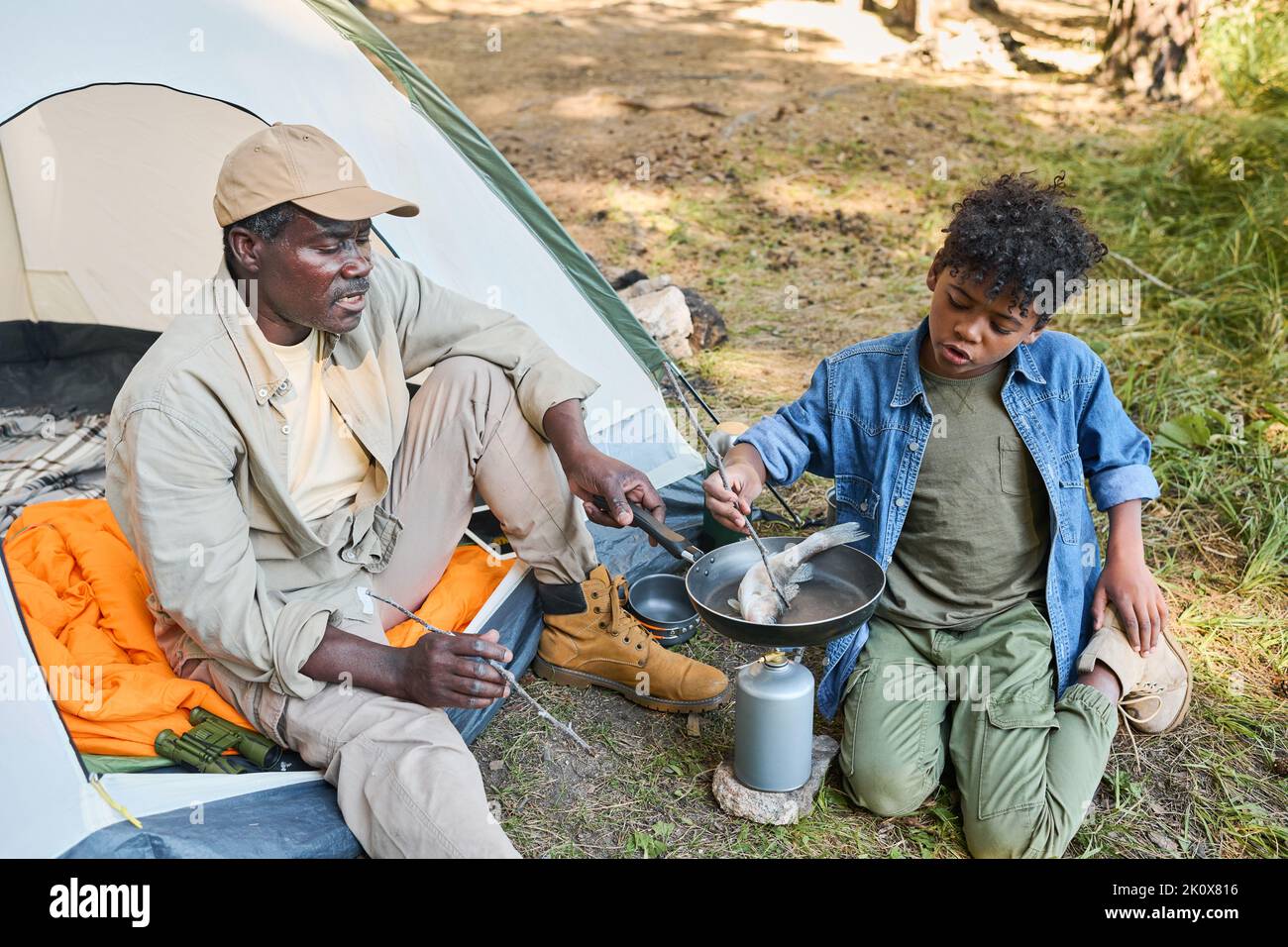 Älterer schwarzer Mann und sein Enkel kochen frischen Fisch auf der Bratpfanne, während sie am Zelt auf dem Boden sitzen und eine Wanderung im Wald genießen Stockfoto