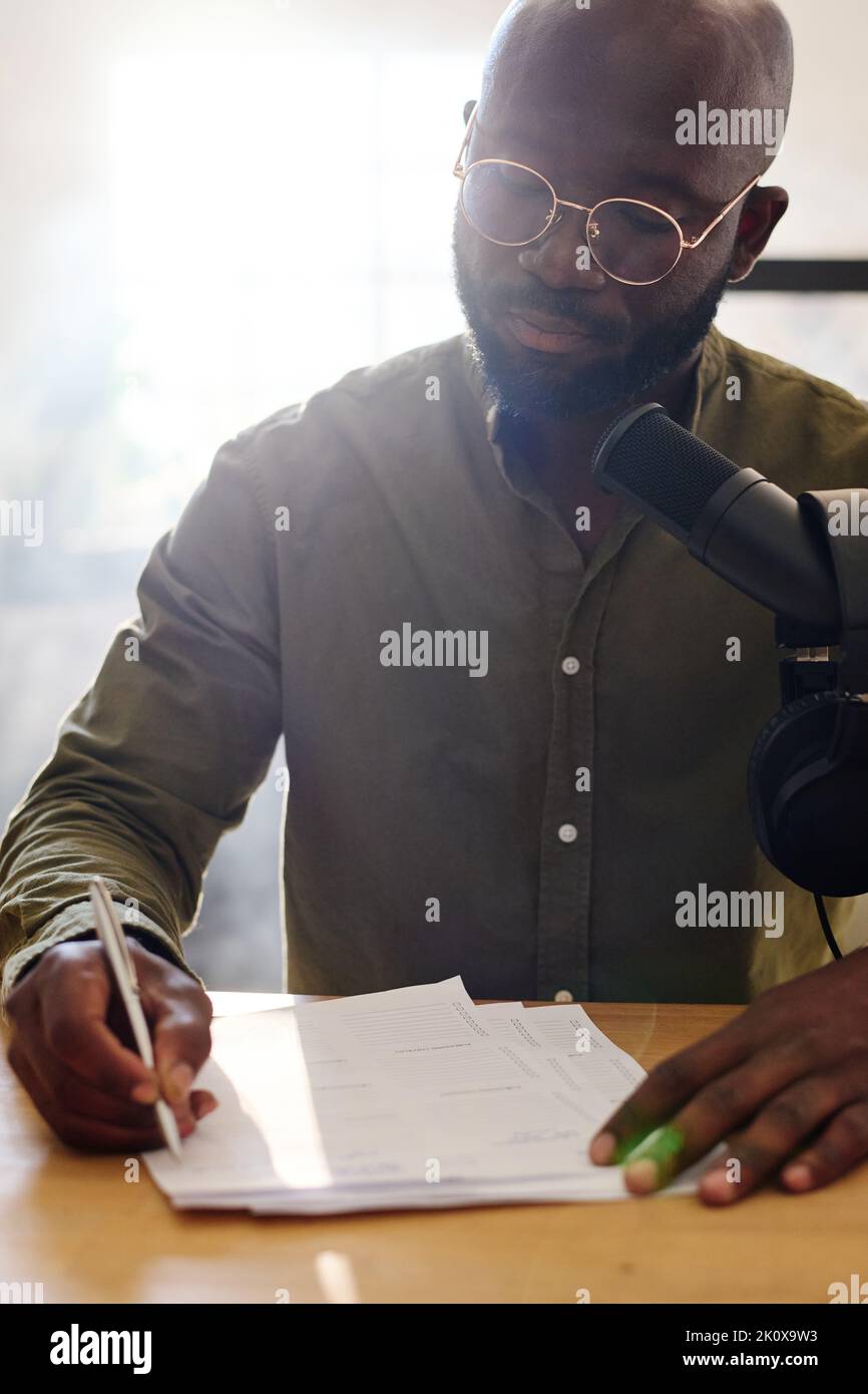 Ein junger, glatzköpfiger schwarzer Mann, der Fragen auf Papier las, die für ein Interview mit einem Gast vorbereitet wurden, während er vor dem Mikrofon am Schreibtisch im Studio saß Stockfoto