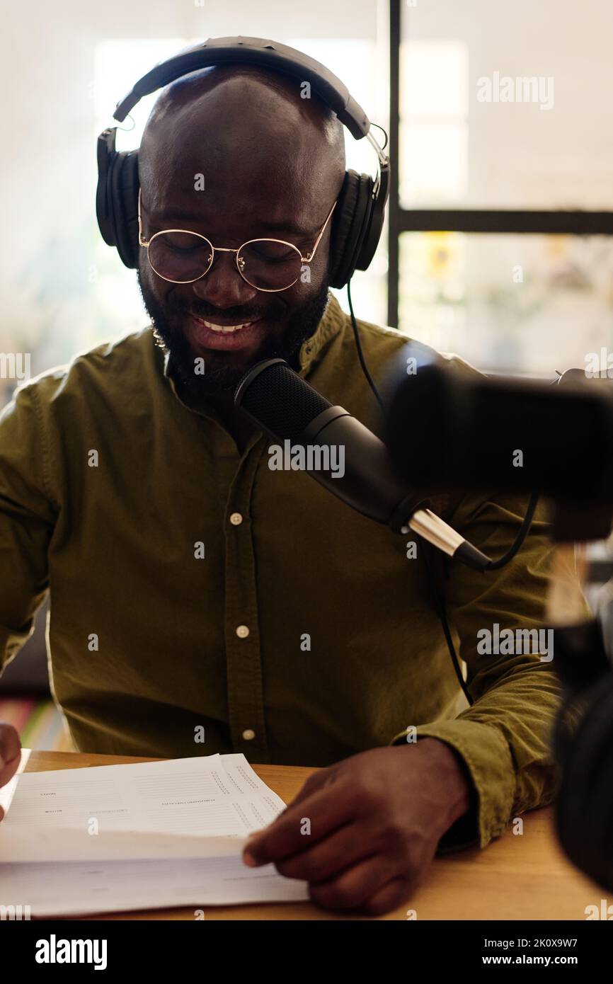 Junger lächelnder afroamerikanischer Mann in Kopfhörern, Brillen und Casualwear, der vor dem Interview Fragen auf Papier durchschaut Stockfoto