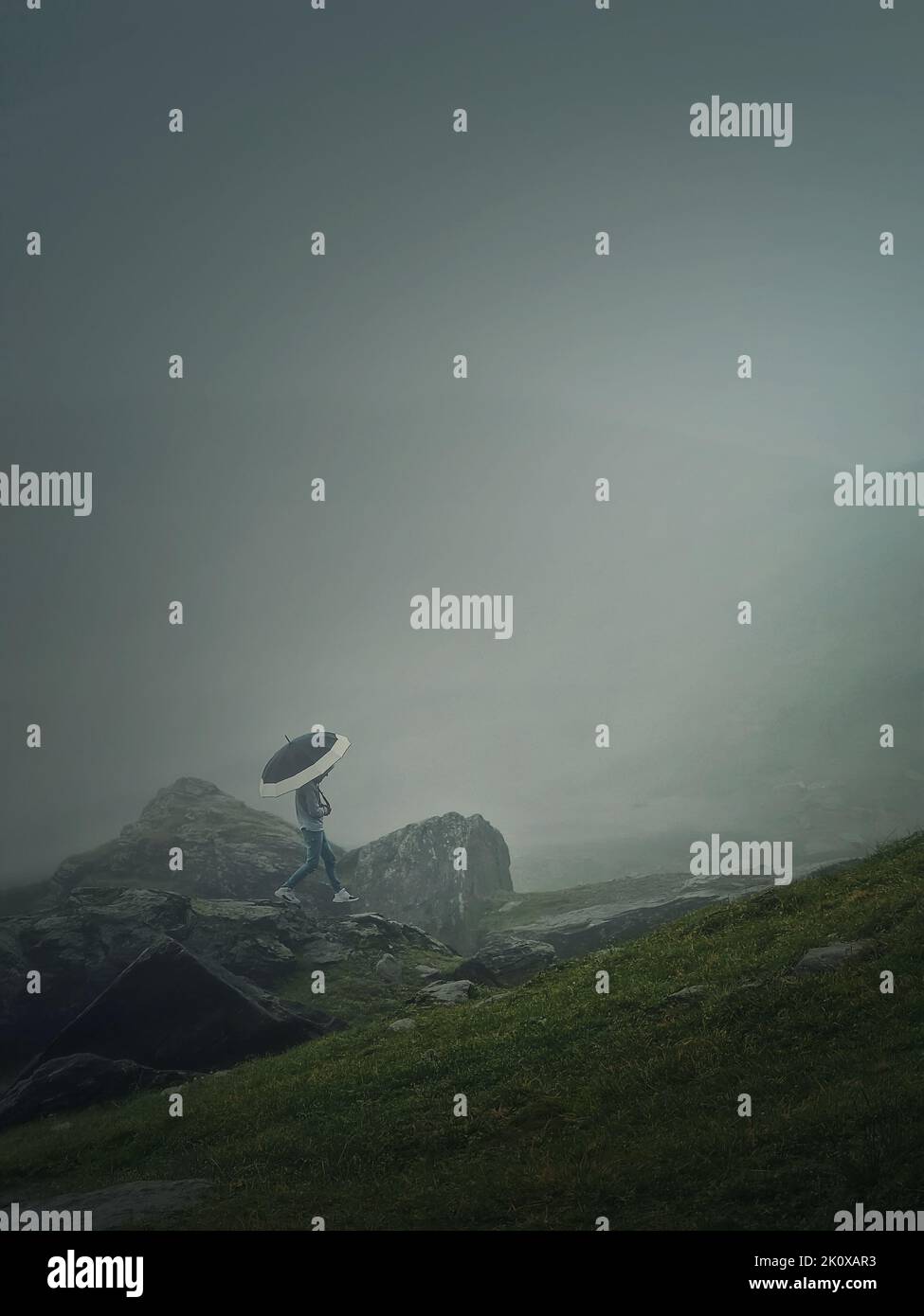 Mann mit Regenschirm, der in einem düsteren Wetter mit dichtem Nebel einen Berg besteigen kann. Moody Wanderszene mit einem Wanderer, der auf den Klippen des nebligen Stockfoto