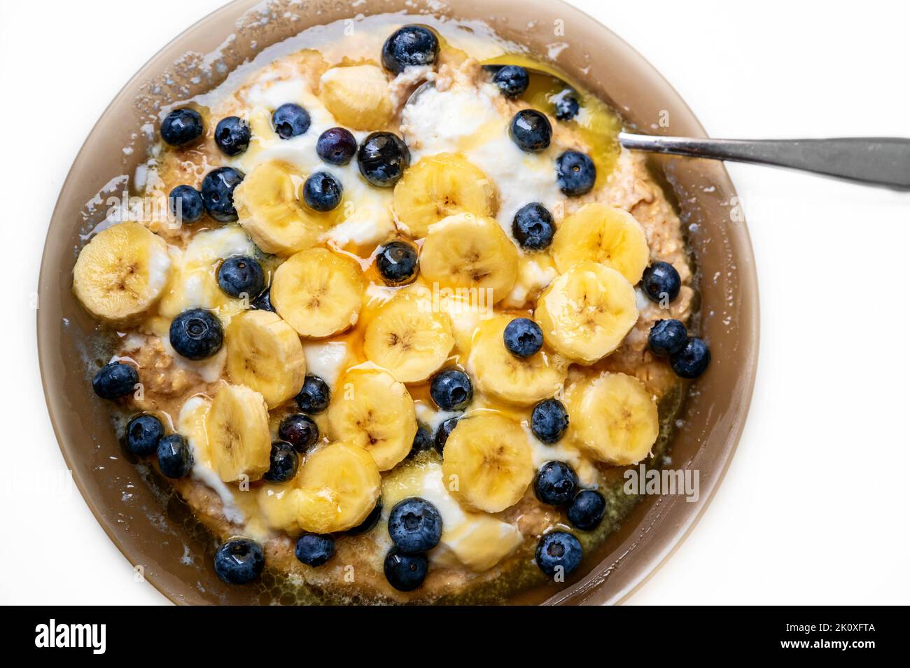 Banane in Kreis geschnitten mit Heidelbeere, Honig und geschmolzener Butter auf Joghurt-Brei auf braunem Teller auf weißem Hintergrund mit Löffel, Nahaufnahme. Süß Stockfoto