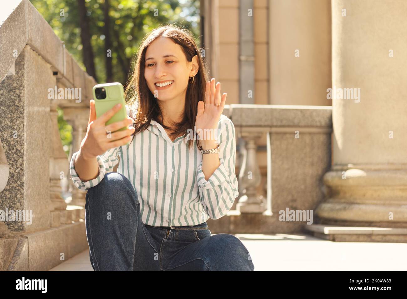 Eine junge lächelnde Frau telefoniert mit einem Videoanruf, den sie an einem sonnigen Tag auf einer Treppe anruft. Stockfoto