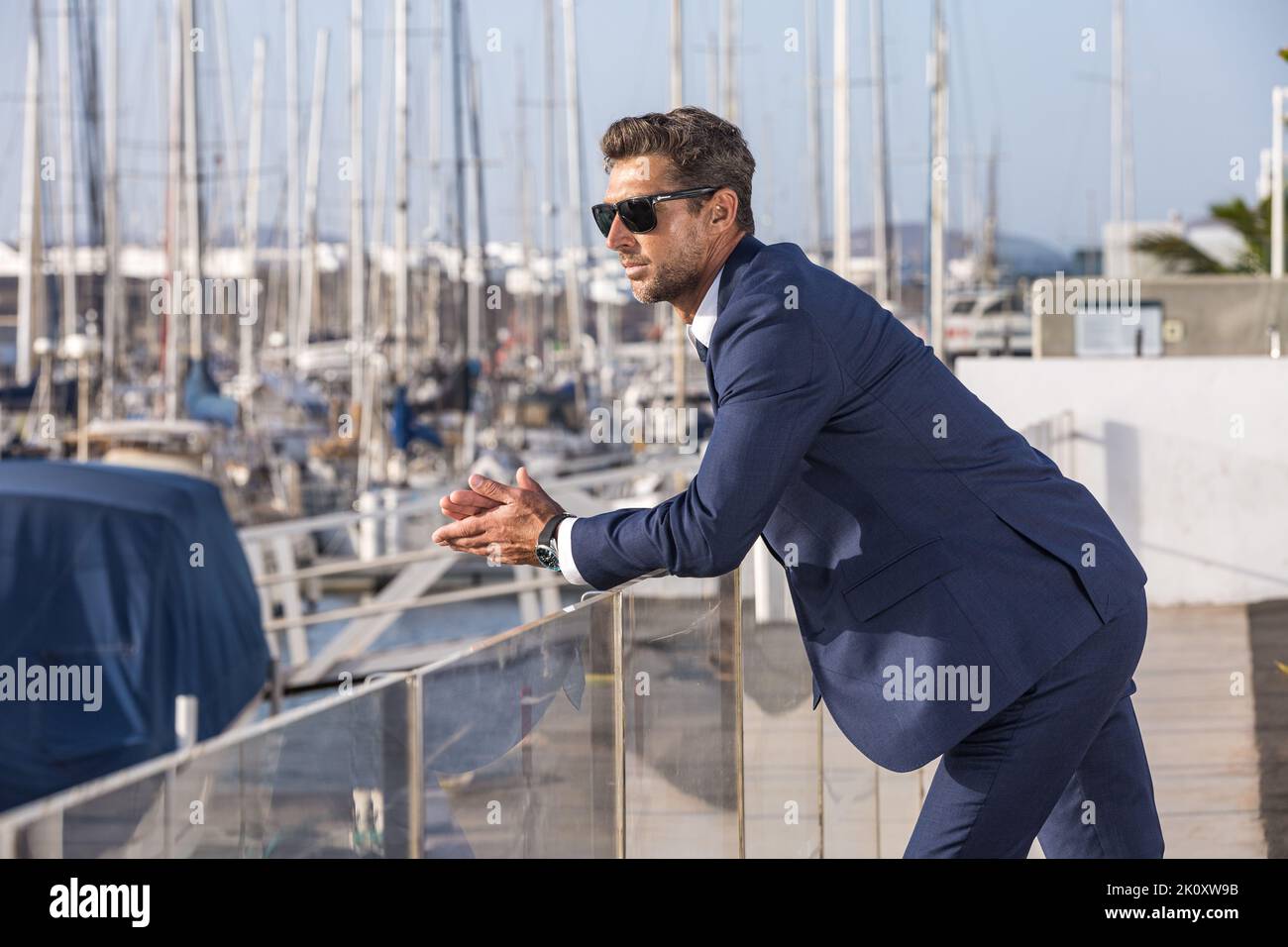 Seitenansicht eines männlichen Unternehmers in Anzug und Sonnenbrille, die am Glaszaun gelehnt ist und das Meerwasser auf unscharfem Hintergrund von Yachten an sonnigen Tagen in CI bewundert Stockfoto