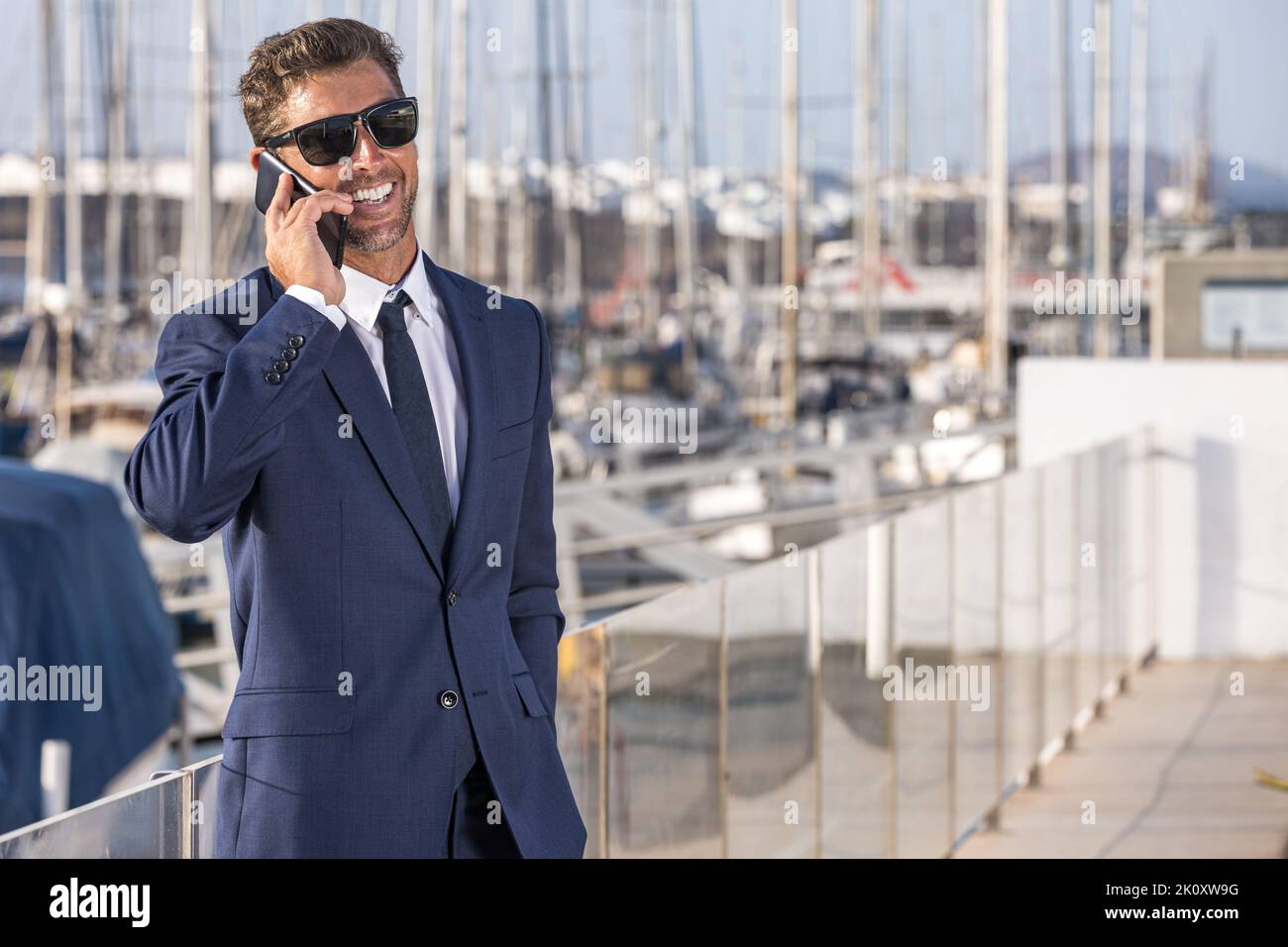 Fröhlicher Geschäftsmann in Anzug und Sonnenbrille lächelt und redet auf dem Smartphone, während er einen sonnigen Sommertag im Stadthafen verbringt. Stockfoto