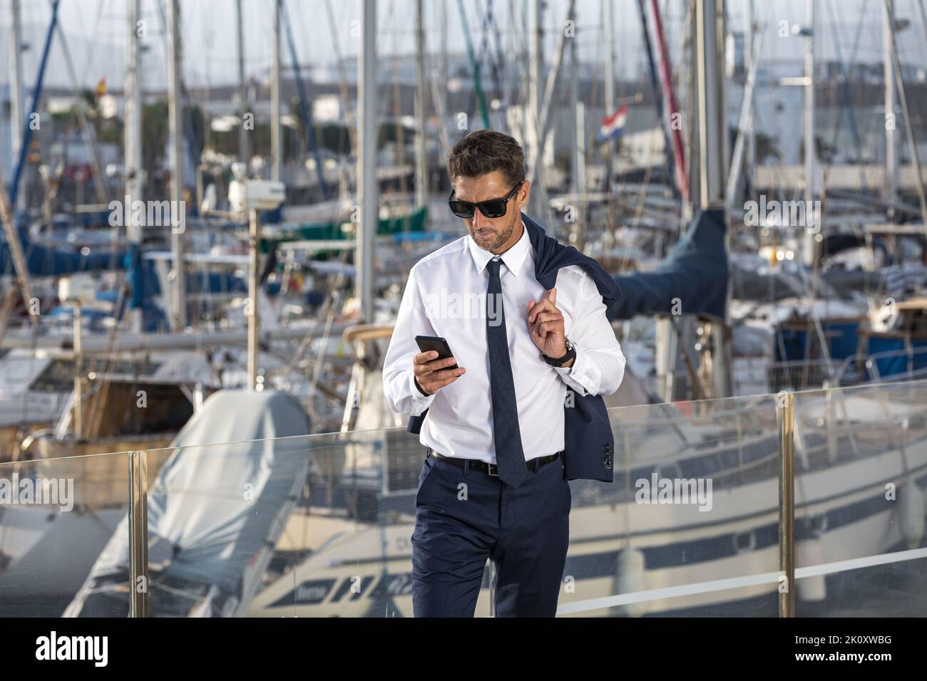 Männlicher Unternehmer in formeller Kleidung und Sonnenbrille mit Jacke auf der Schulter und SMS auf dem Handy, während er auf verschwommenem Hintergrund von Segelbooten steht Stockfoto