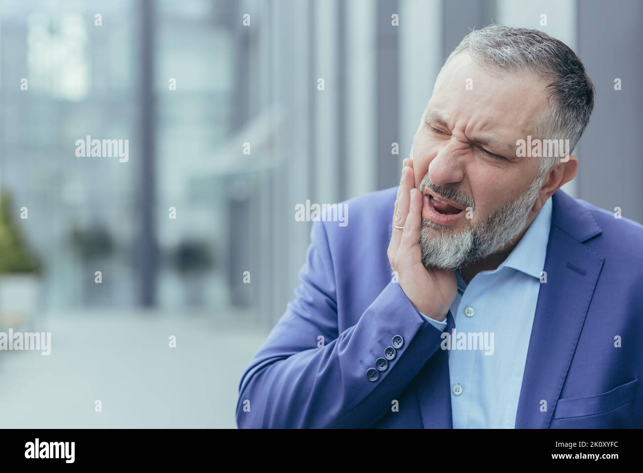 Zahnschmerzen. Nahaufnahme. Älterer Mann, Geschäftsmann, Büroangestellter, der auf der Straße in der Nähe der Arbeit steht, seine Wange hält, unerträgliche Zahnschmerzen empfindet, sich grimazend anfühlt, braucht Hilfe. Stockfoto