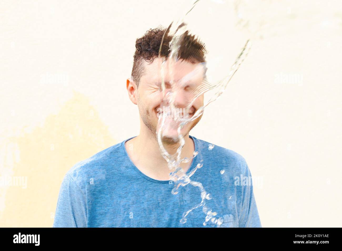 Nass lächelnder junger kaukasischer Mann in blauem T-Shirt mit geschlossenen Augen im Wasserspritzer, Wasserstrahl auf gelbem Hintergrund. Stress, Schock. Vorderansicht, Port Stockfoto