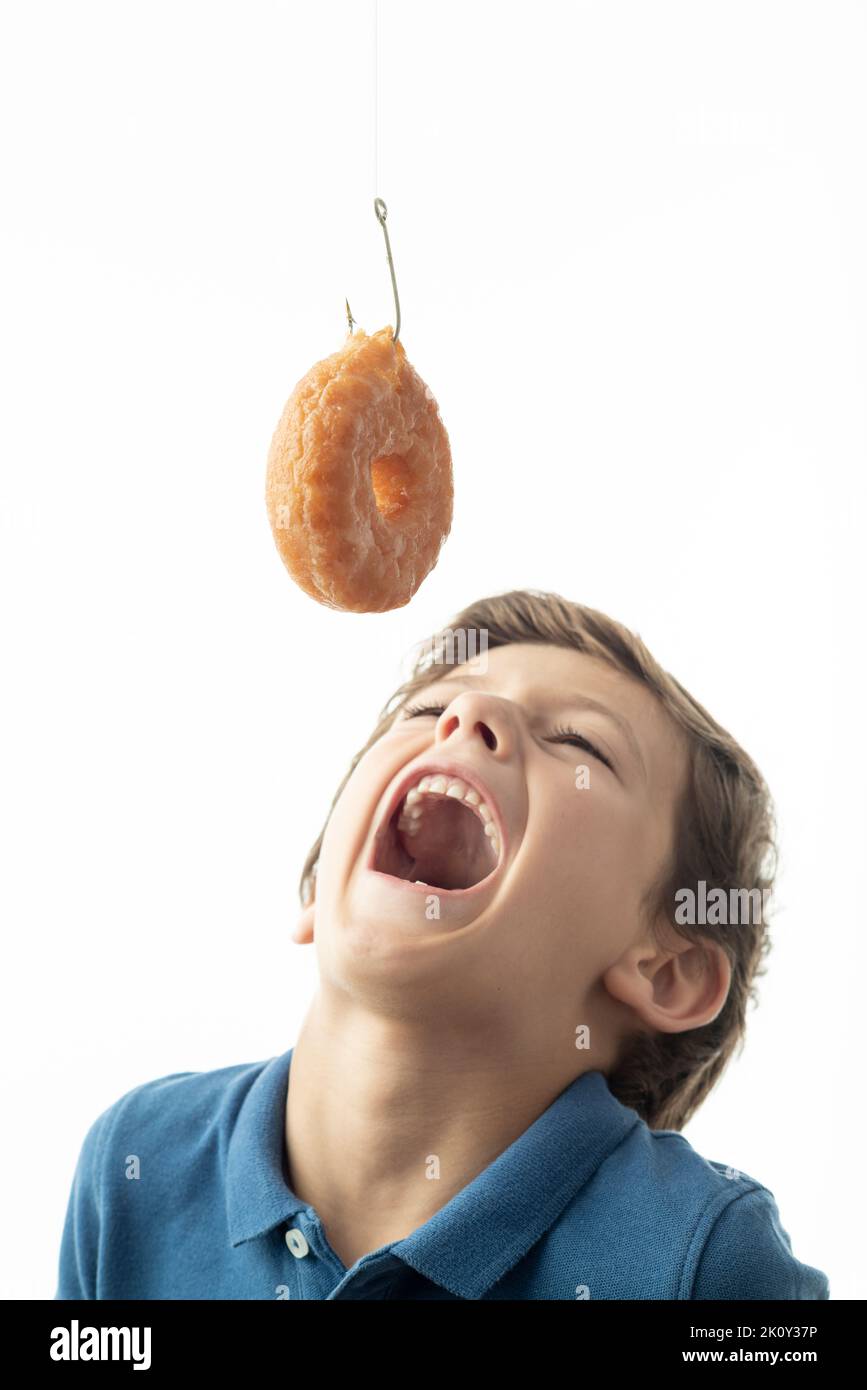 Ein Kind versucht, einen Donut, der an einem Haken hängt, zu beißen. Das Bild zeigt das Konzept der Suchtproblematik gegen Zucker und süße Lebensmittel. Weißer Hintergrund und Platz für Stockfoto