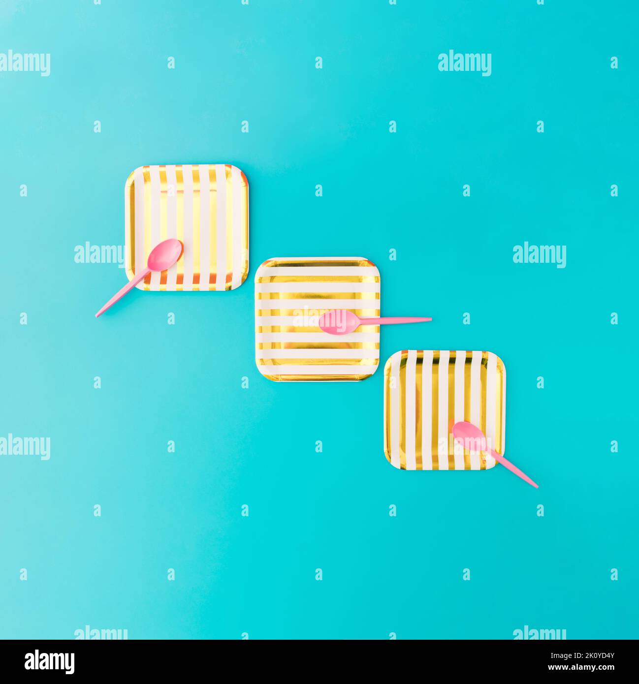 Picknick-Set mit drei Gold- und weiß gestreiften Papptellern und drei pinken Plastiklöffeln auf blauem Hintergrund. Minimalkonzept. Stockfoto