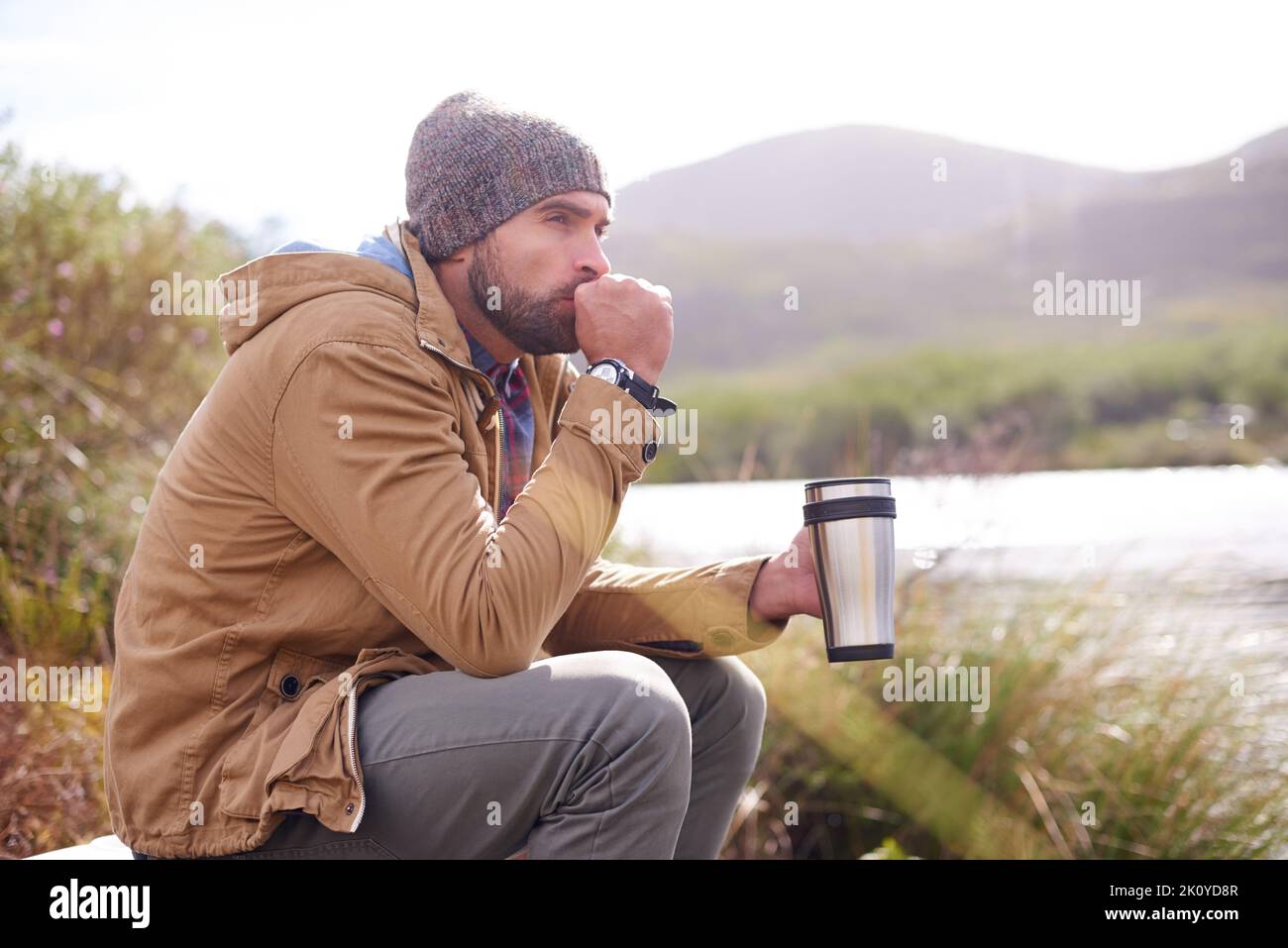 Genießen Sie einen knackigen Morgen am See. Ein hübscher Mann, der mit einer Thermoskanne an einem See sitzt. Stockfoto