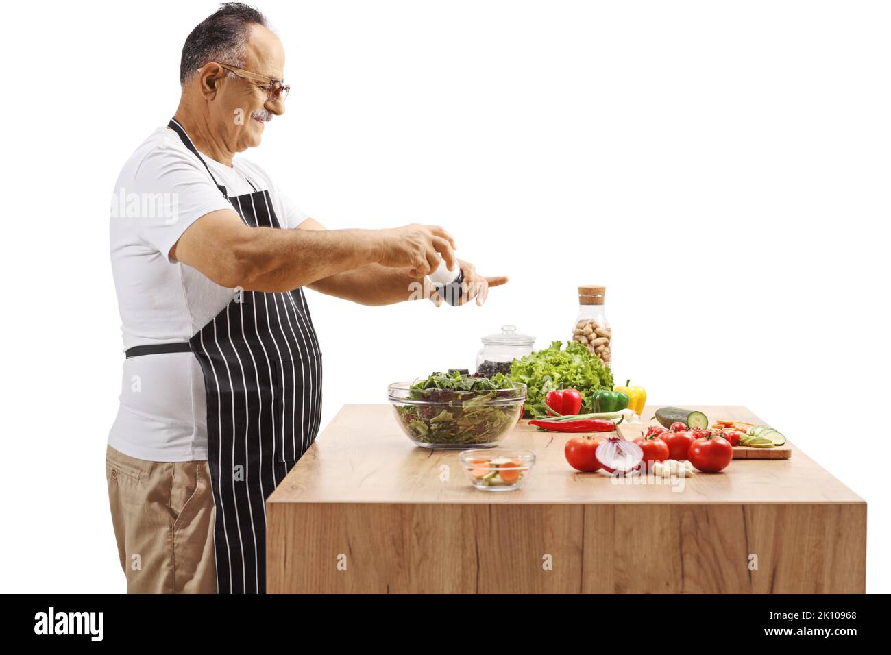 Seitenaufnahme eines reifen Mannes, der einen Salat auf einer Küchentheke würzt, die auf weißem Hintergrund isoliert ist Stockfoto