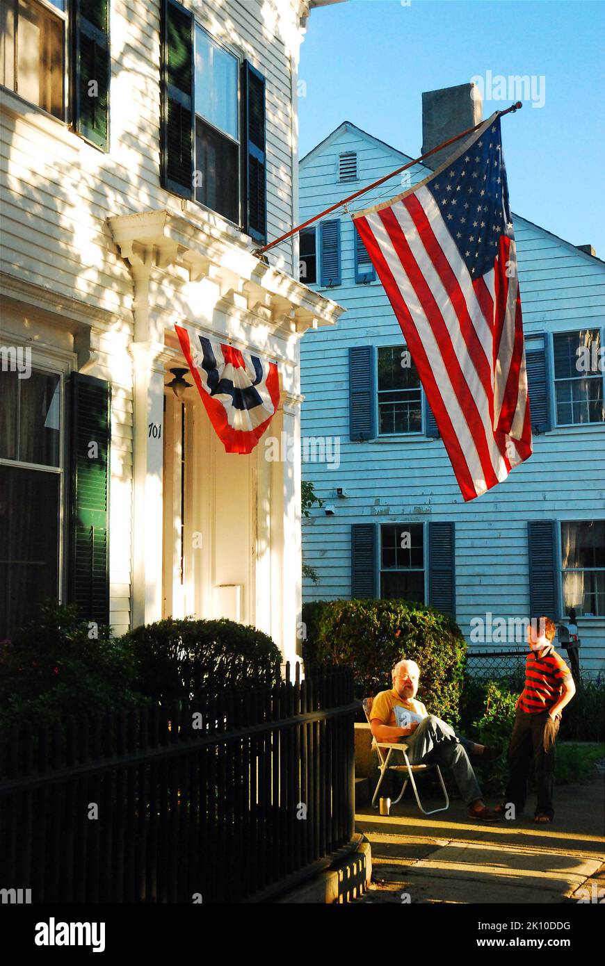 Ein Großvater und sein Enkel unterhalten sich am 4. Juli unter amerikanischer Flagge im Vorgarten ihres Hauses. Stockfoto