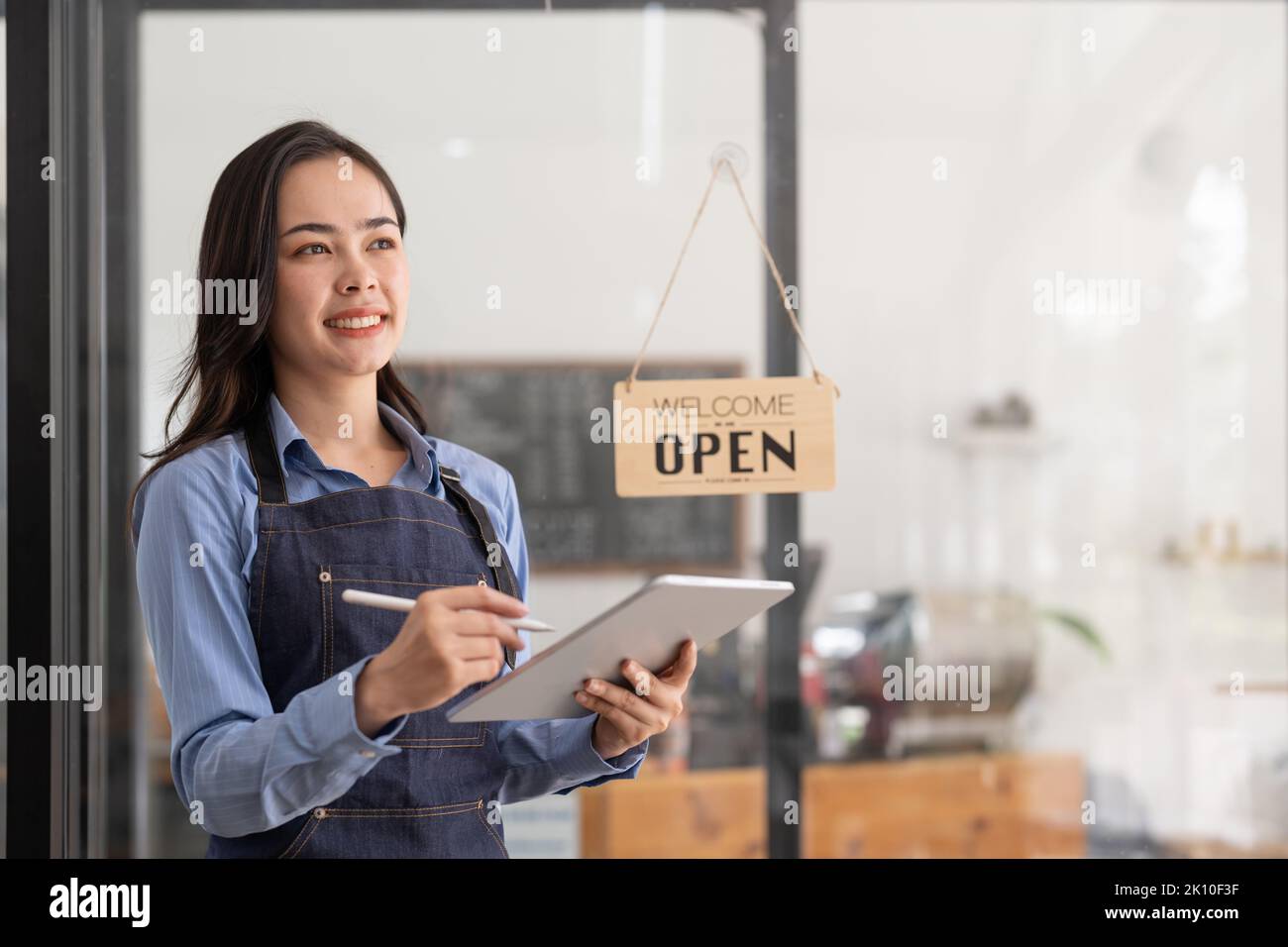 Schöne asiatische junge Barista Frau in Schürze hält Tablet und vor der Tür des Cafés mit offenem Schild stehen. Start des Geschäftsinhabers Stockfoto