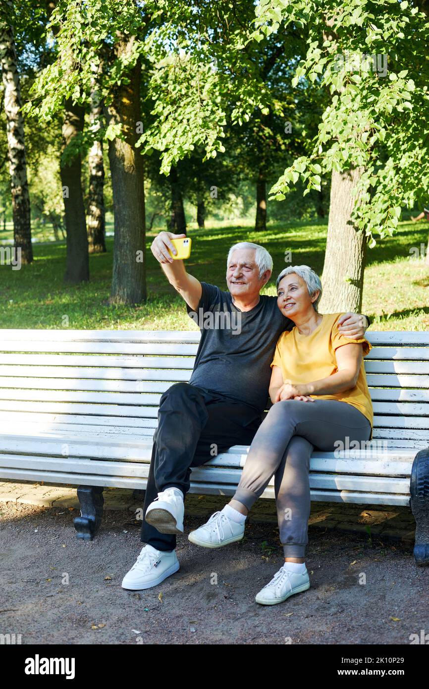 Alte Ehepartner machen Selfie-Bild oder Videoanruf sitzen auf der Bank im Sommerpark. Grauhaarige Paar Blick auf Handy-Bildschirm machen Videocall im Freien verwenden mod Stockfoto