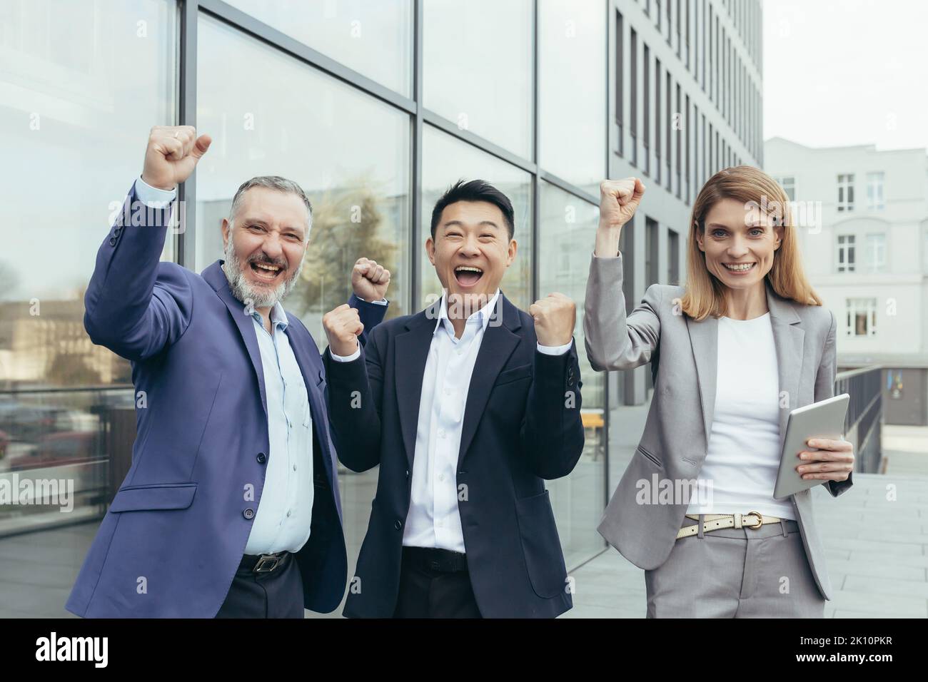 Interrassische Geschäftspartnerschaft. Drei Geschäftsleute feiern einen Sieg, einen erfolgreichen Deal. Ein älterer Mann, ein junger asiatischer Mann und eine junge Frau in Anzügen in der Nähe des Büros, lachen, zeigen mit ihren Händen ja. Stockfoto