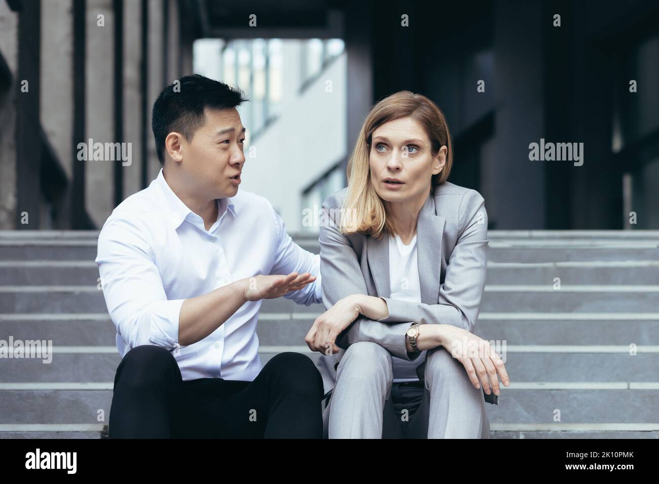 Interrassischer Konflikt am Arbeitsplatz. Eine junge Frau und ein junger asiatischer Mann sitzen auf den Stufen vor einem Büro und streiten sich. Ein Mann schreit und beweist einer Frau etwas. Die Frau hört erittlich zu. Stockfoto