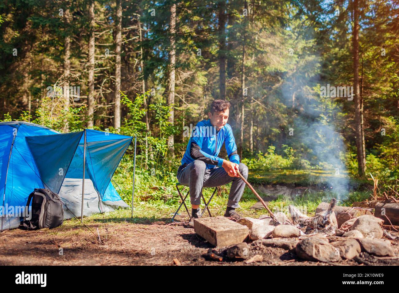 Ein Mann, der sich am Lagerfeuer im Wald entspannt, sitzt neben dem Zelt und kontrolliert das Feuer. Camping im Sommer. Allein reisen und die Natur genießen Stockfoto