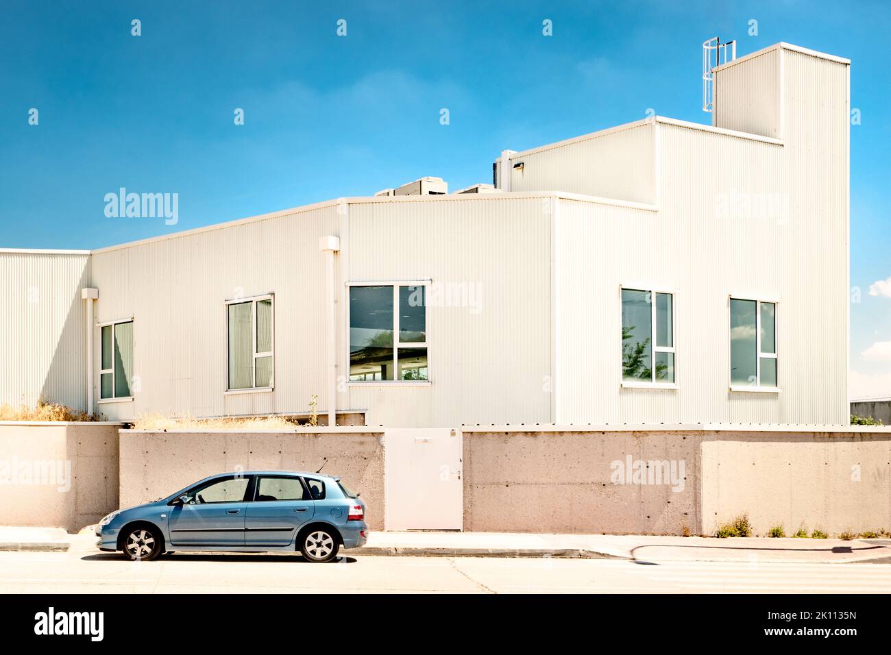 Fassade eines Industrielagers mit weißem Dach, Zementwand, Fenstern und geparktem Auto an einem sonnigen Tag Stockfoto