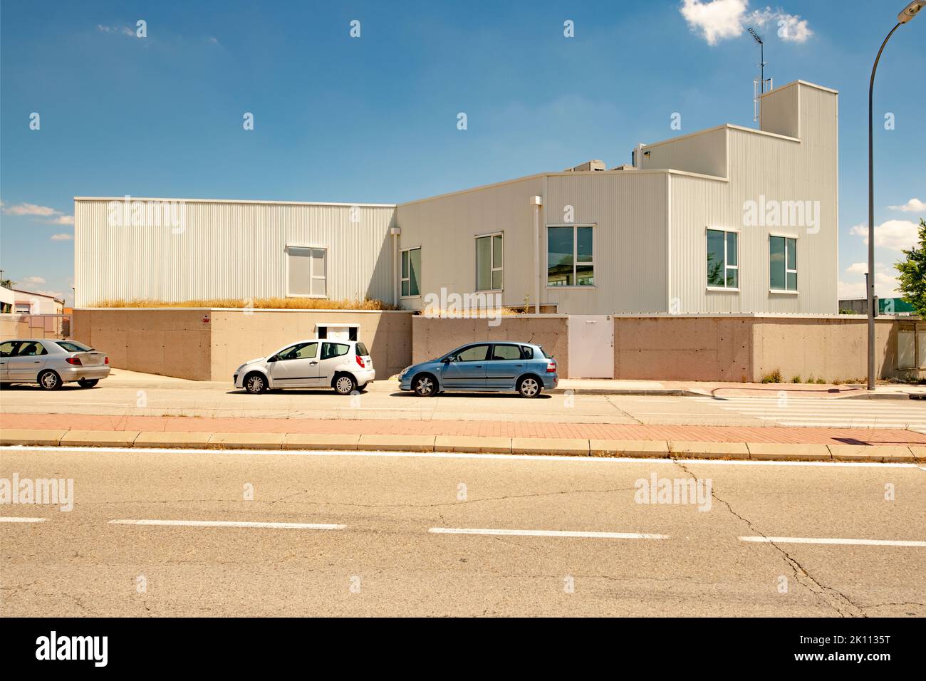 Fassade eines Industrielagers mit weißem Dach, Zementwand, Fenstern und geparkten Privatautos an einem sonnigen Tag Stockfoto