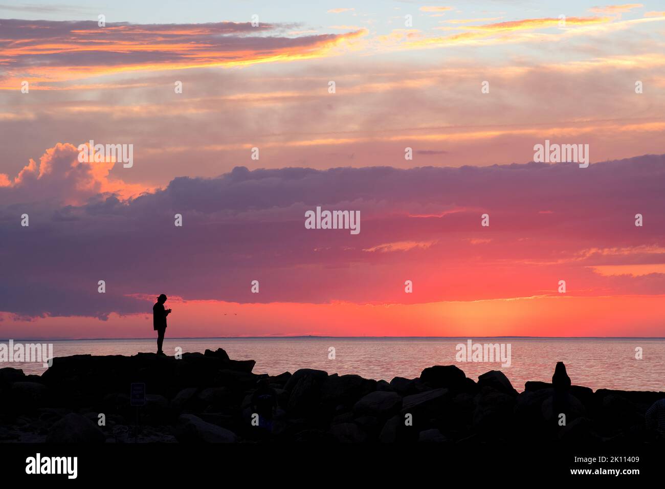 Dramatische Szene an der Küste von Neuengland. Dunkle Silhouetten zweier unverkennbarer Menschen auf Wellenfelsen am Cape Cod Beach, die den farbenfrohen Sonnenuntergang betrachten. Stockfoto