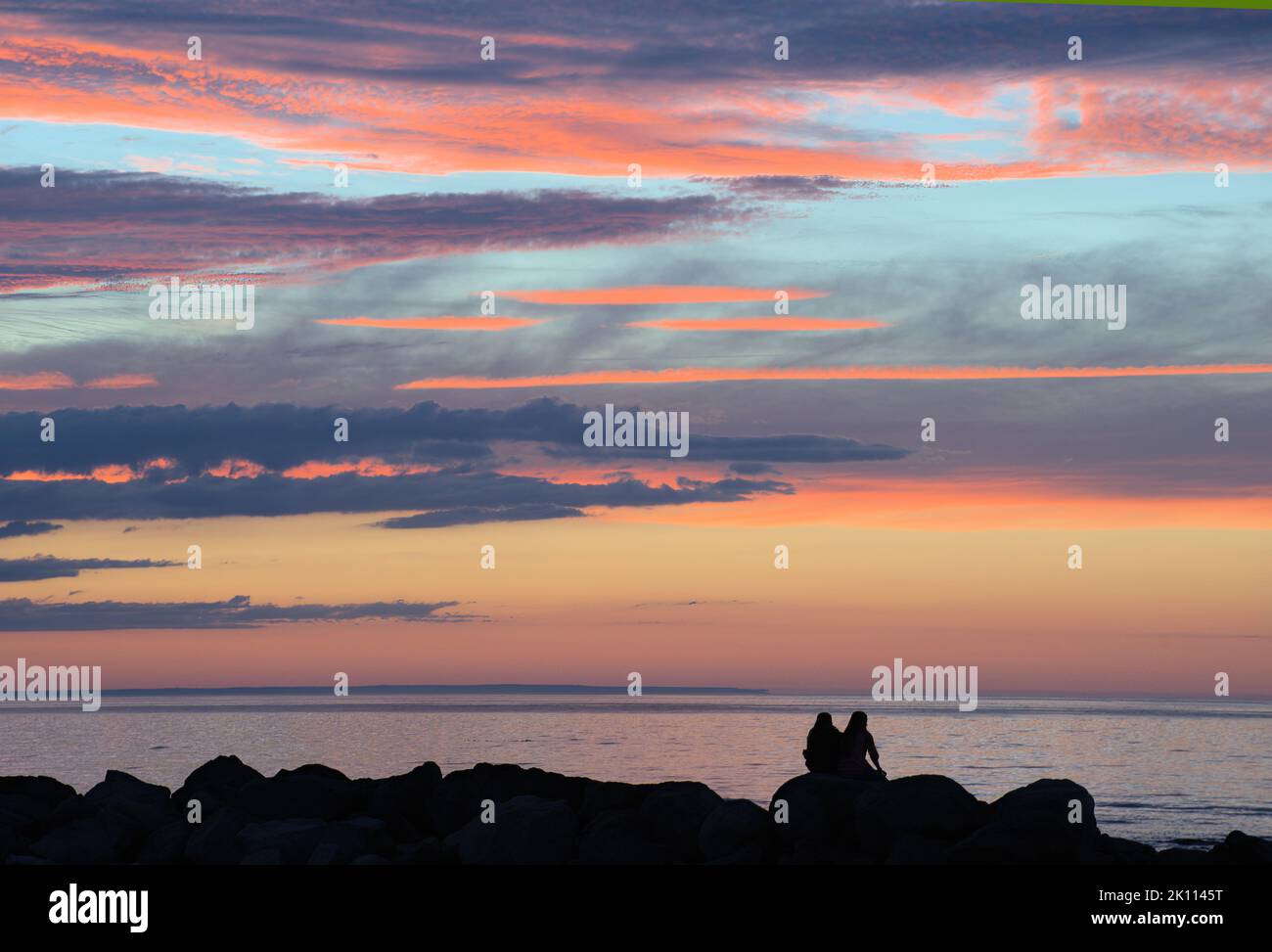 Ruhige Dämmerungsszene am Cape Cod Beach. Dunkle Silhouetten zweier unkenntlich gegannter Frauen, die auf Wellenfelsen sitzen und den farbenfrohen Sonnenuntergang betrachten. Stockfoto