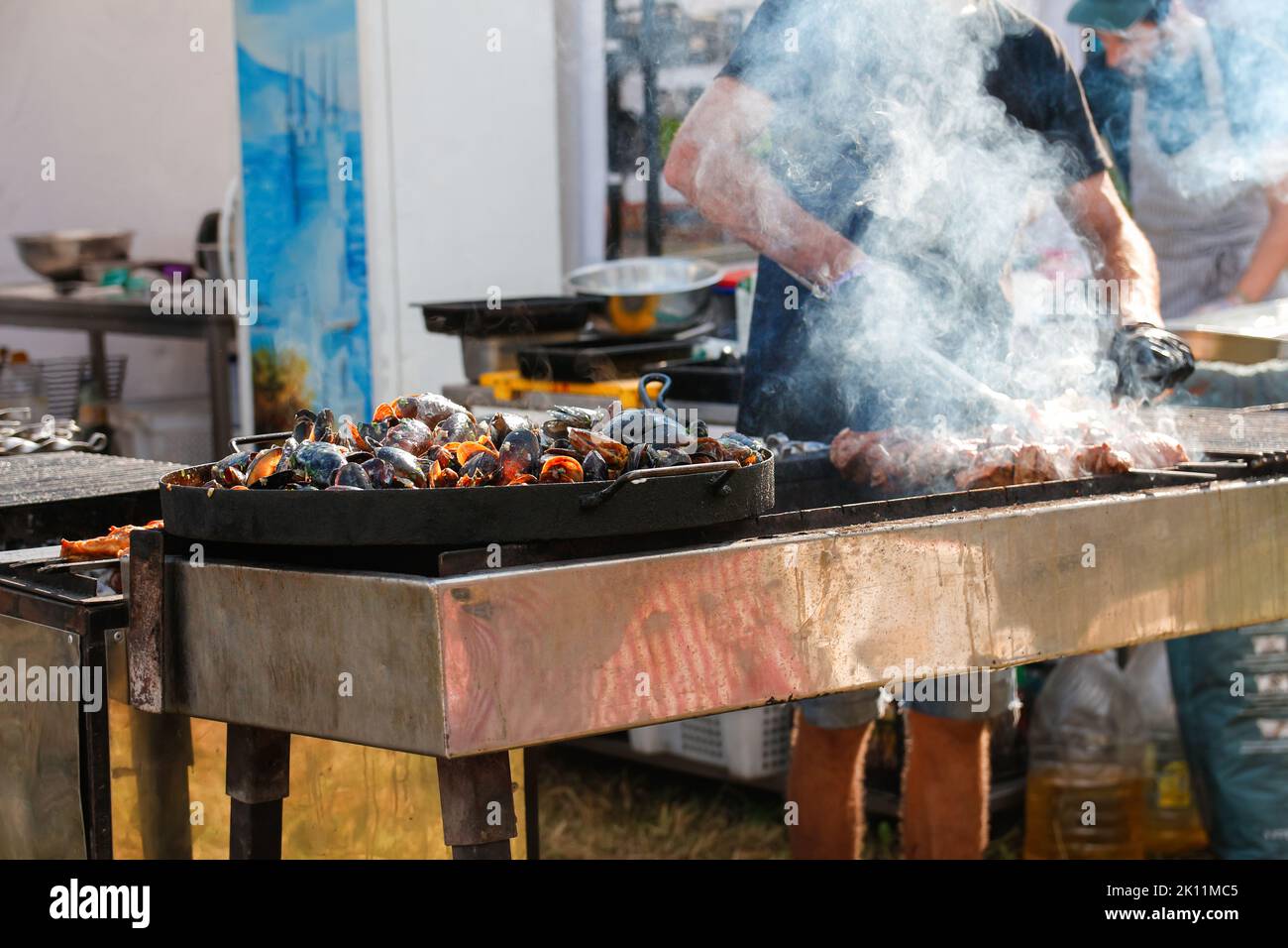 Unschärfe Mann Kochen bbq Fleisch auf Festival im Freien. Paella mit Meeresfrüchten. Der Koch gegrillten Würstchen im Park draußen. Konzept der Sommerparty mit Familien und Stockfoto
