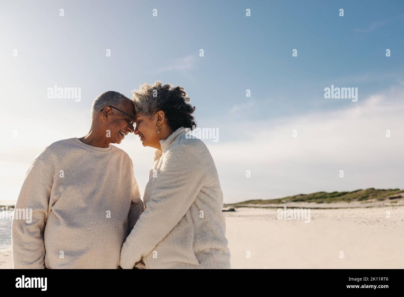 Liebevolles Seniorenpaar lächelt und berührt am Strand ihre Köpfe zusammen. Reifes Paar, das einen romantischen Moment im Freien teilt. Fröhliche ältere Leute Stockfoto